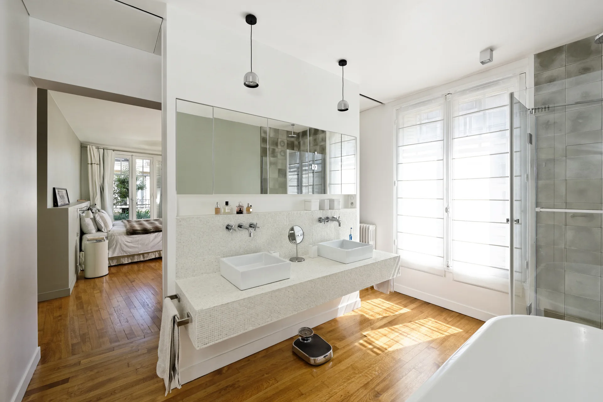 A modern bathroom features a floating double-sink vanity finished with white mosaic tile, hardwood floors, and an adjacent view into the bedroom.