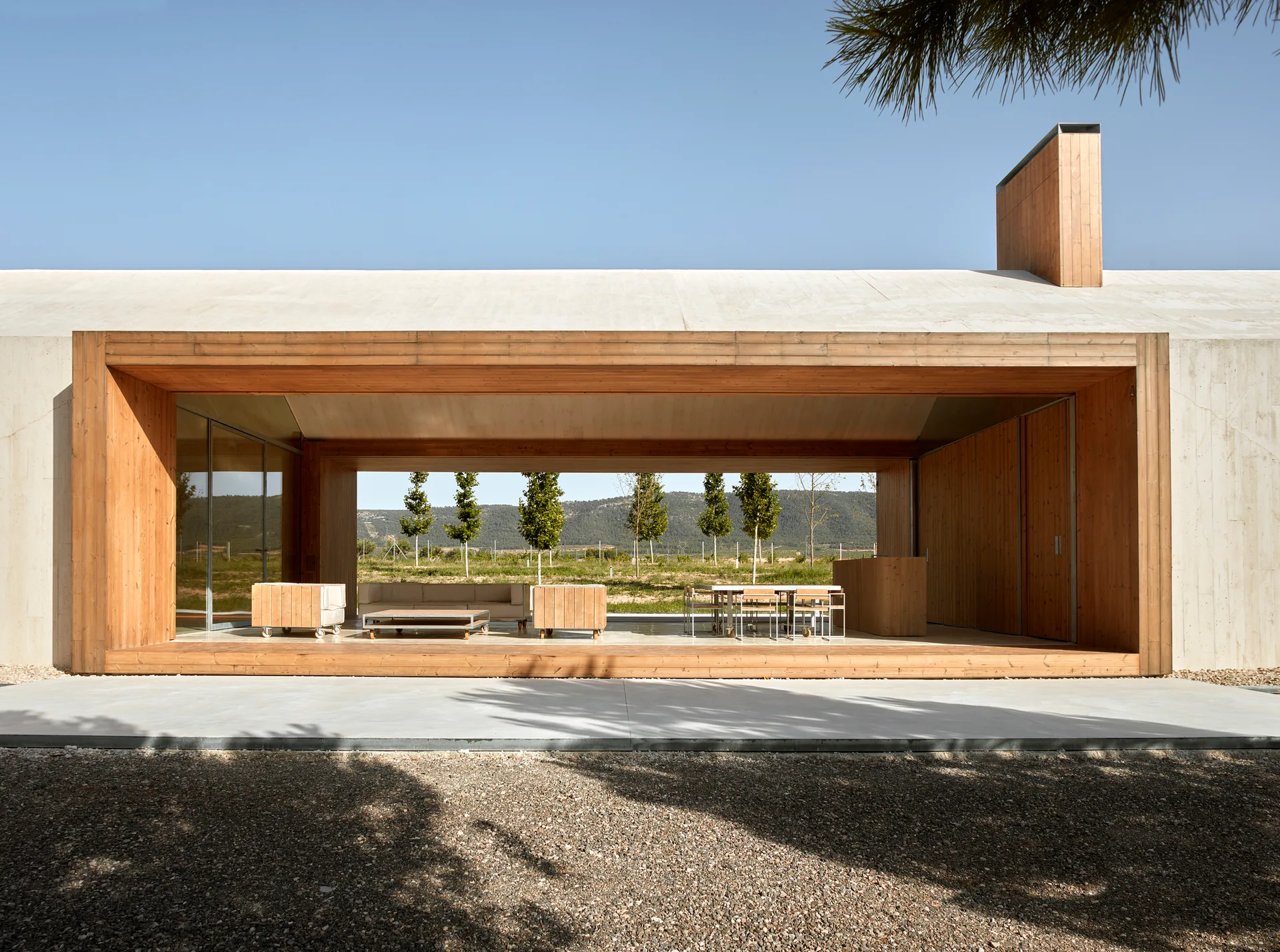 A modern open-air concrete pavilion features a wood-lined interior containing a minimalist lounge area and dining set looking out to a grassy landscape.