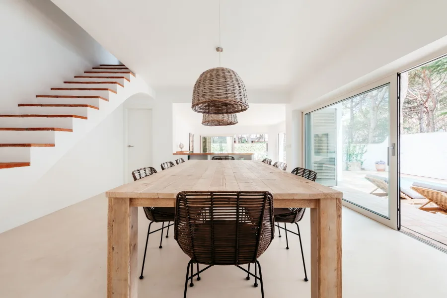 A light wood dining table with dark woven chairs is positioned beneath wicker dome pendant lights in a bright room with a floating wooden staircase.