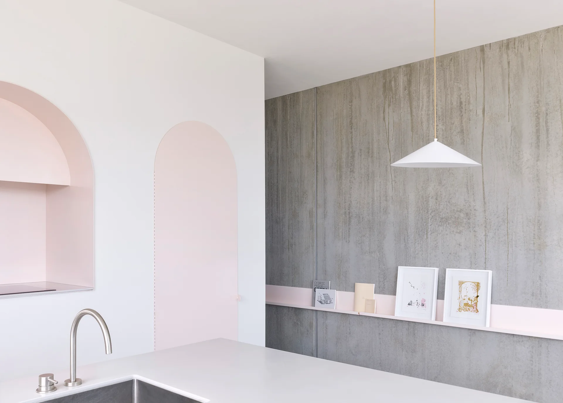 A minimalist kitchen features a white countertop with a sink, pale pink arched wall panels, and a concrete-textured wall with a pink display shelf.