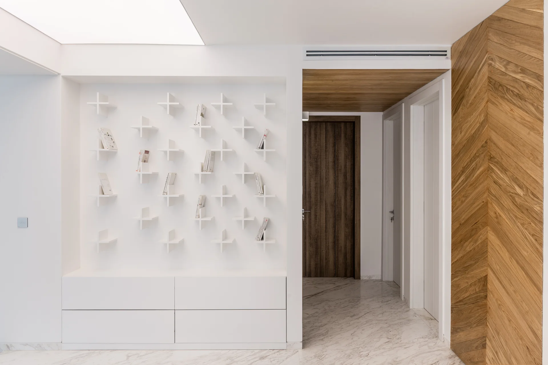 A minimalist white hallway features modular cross-shaped wall shelves over a base cabinet, alongside marble flooring and a chevron-patterned wood wall.
