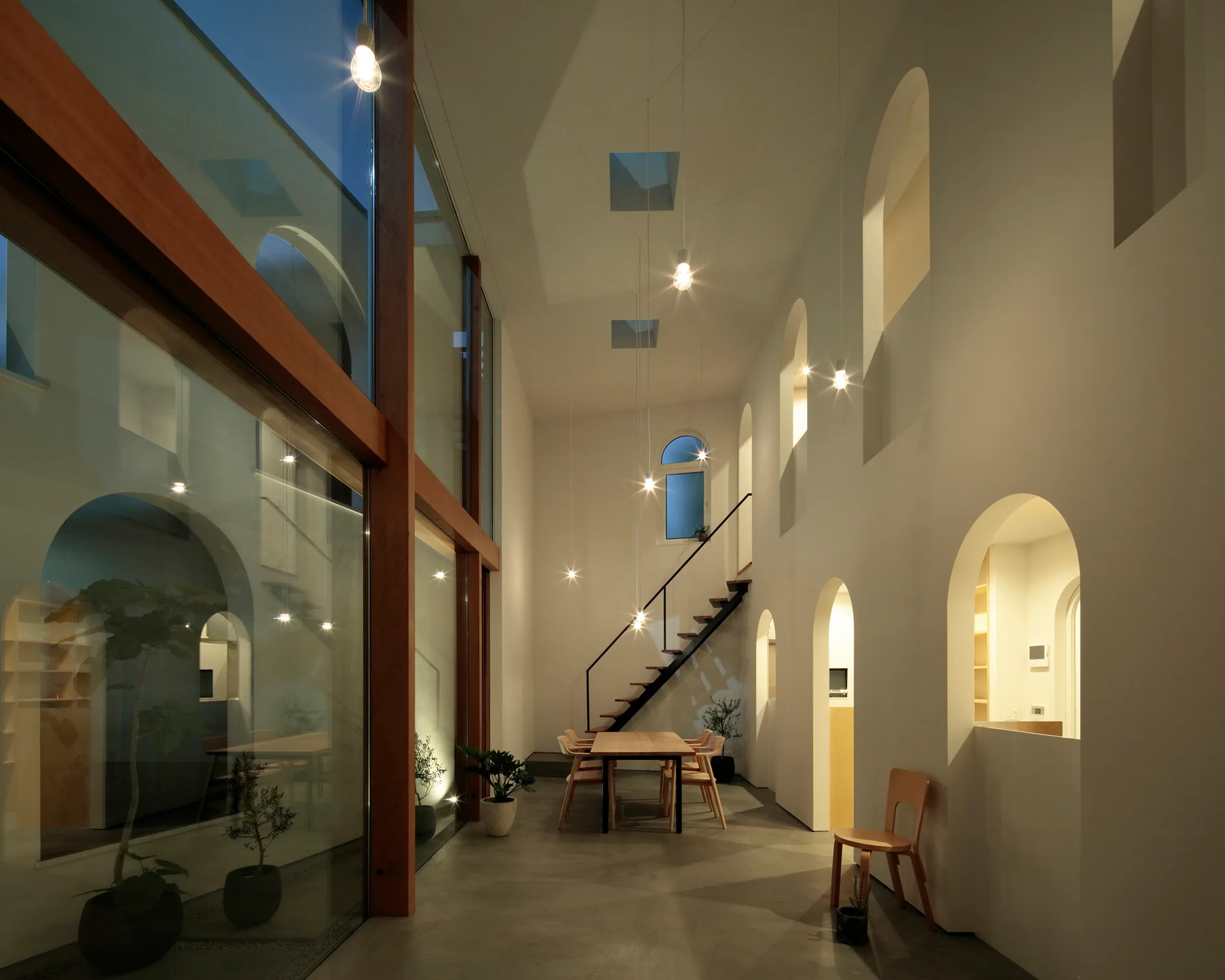A minimal double-height dining room features a wooden table, concrete floors, an open-riser staircase, and a tall wall with multiple arched openings.