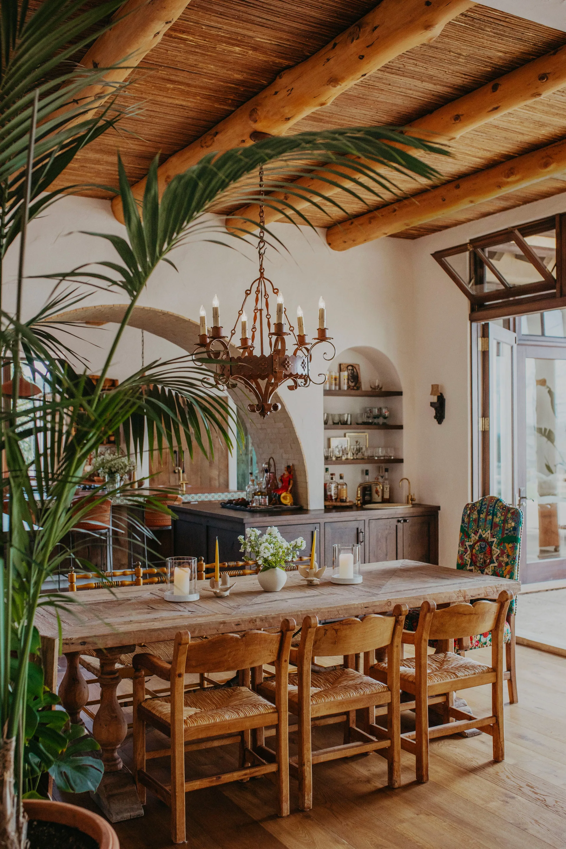 A rustic dining room featuring a long wooden table, chairs with woven seats, an ornate metal chandelier, and a ceiling with exposed log beams.