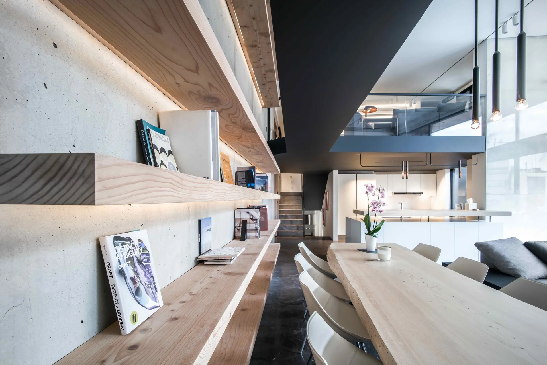 A modern dining room features a long live-edge wood table, white chairs, and floating wood shelves on an exposed concrete wall.