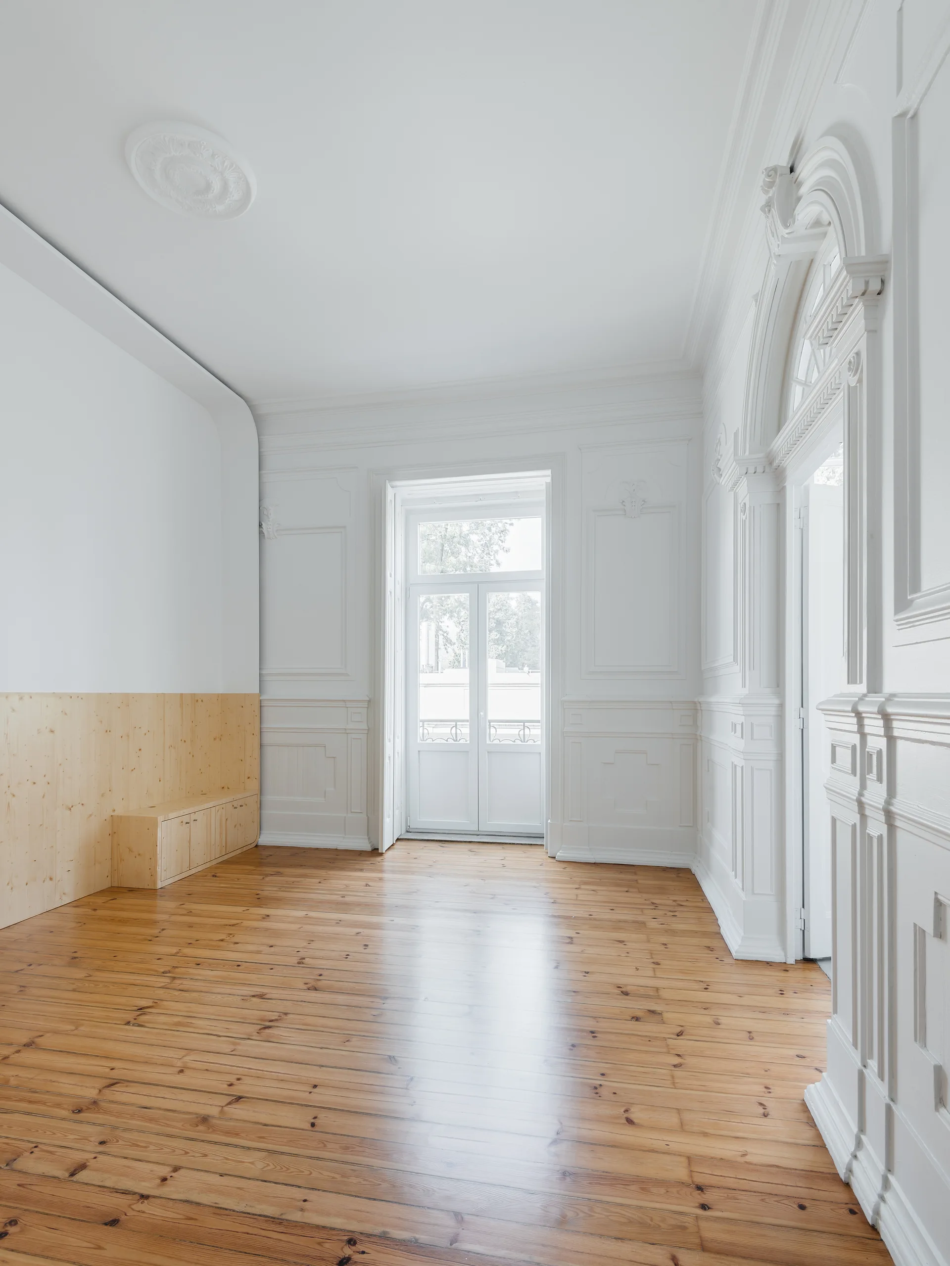 An empty room features white ornate wall moldings, a modern light-wood bench along one wall, and polished wood floors beneath a tall glass door.