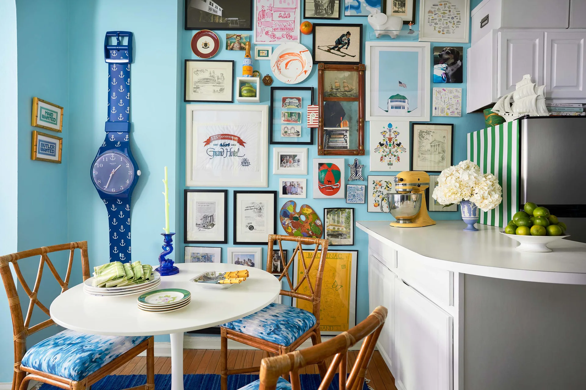 An eclectic dining nook features a white pedestal table, rattan chairs with blue ikat cushions, and a dense gallery wall on bright blue paint.