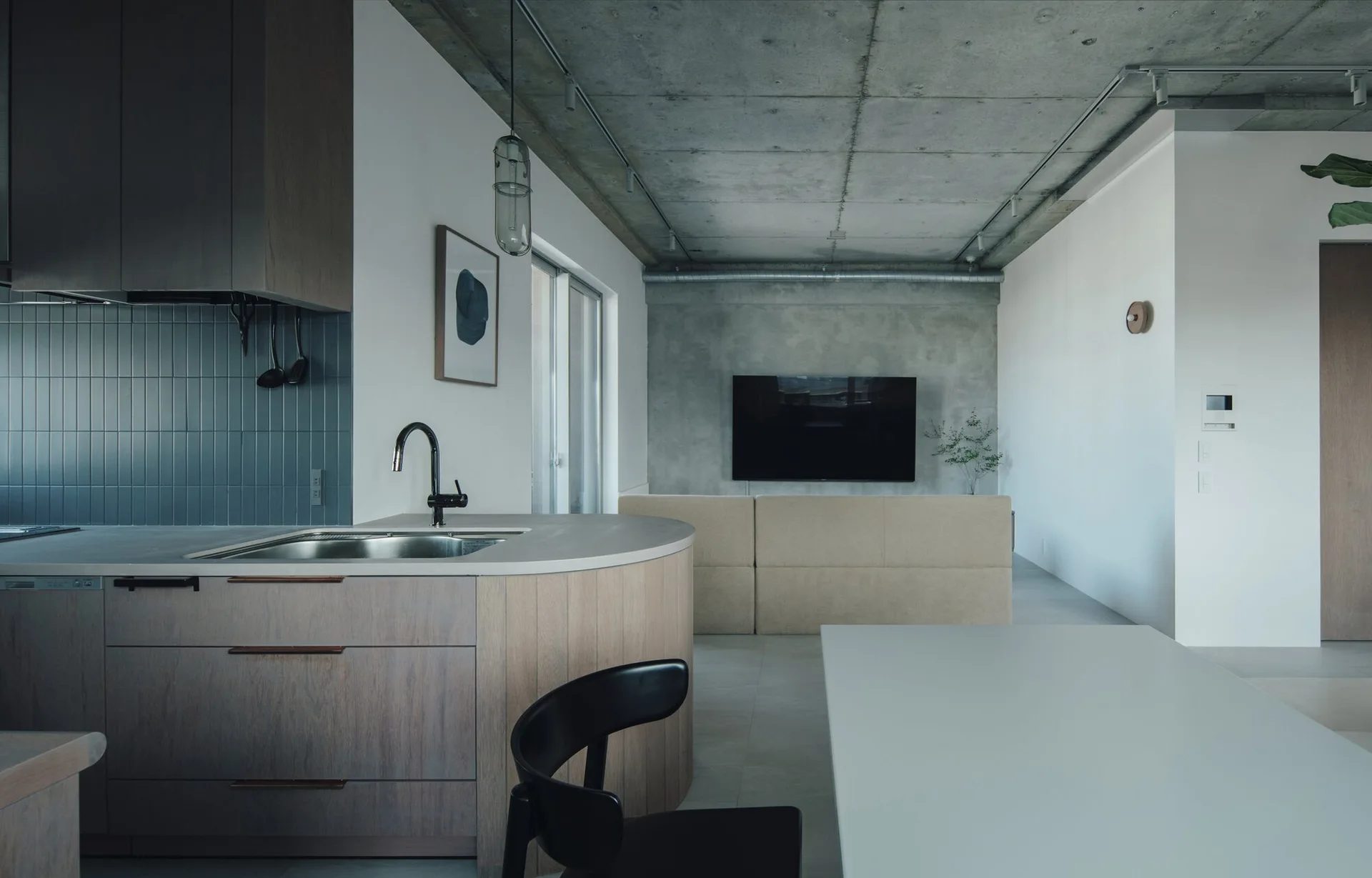 An open-concept kitchen and living area featuring a curved wood kitchen island with a sink, exposed concrete ceilings, and a gray-tiled backsplash.