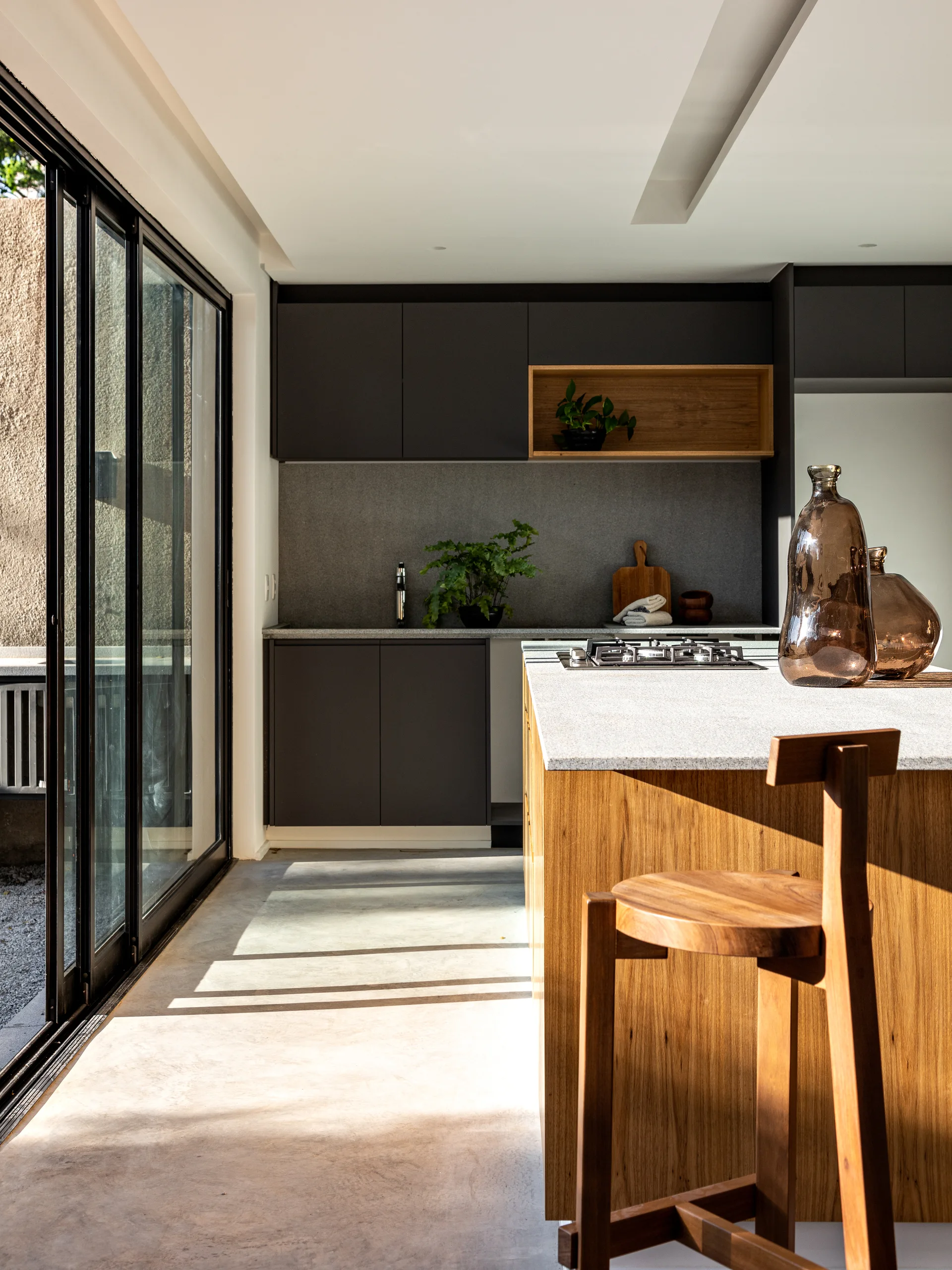 A modern kitchen featuring charcoal matte cabinets, a wooden island with a stone-like countertop, a wooden bar stool, and sliding glass doors.