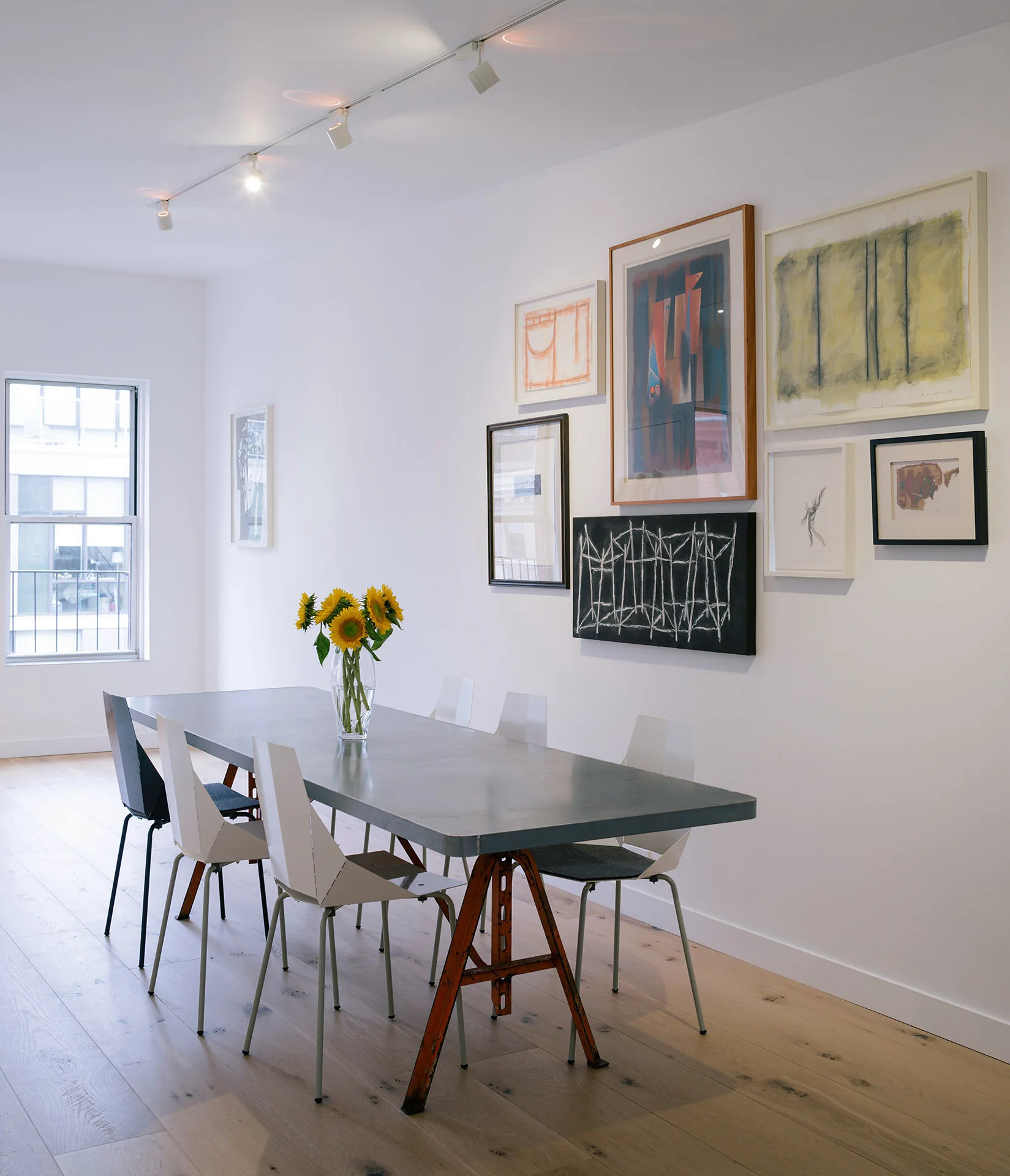 A long gray trestle dining table with modern chairs is set in a white room featuring a gallery wall of framed art and light wood floors.
