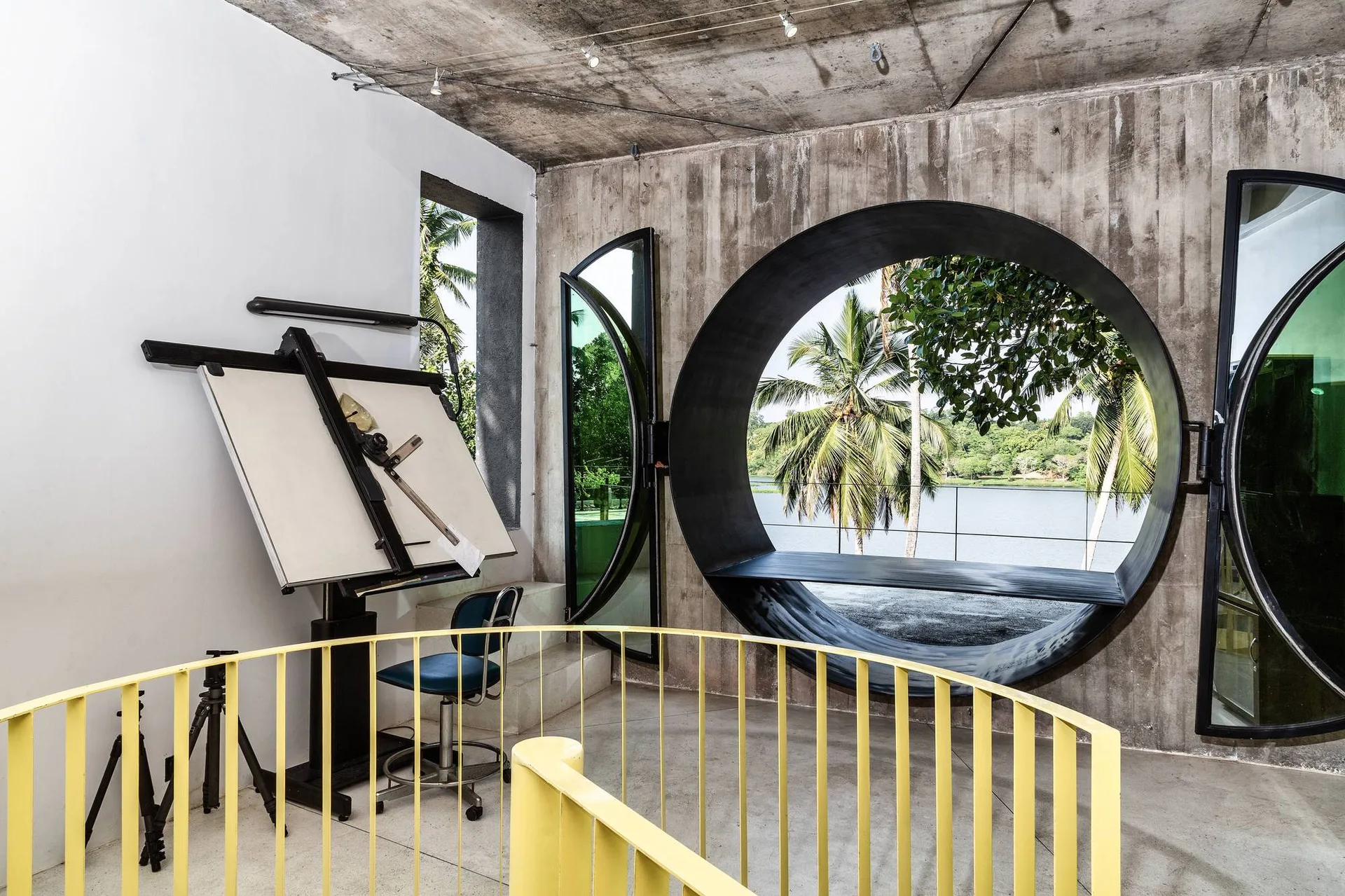 An industrial home studio featuring a large circular window, board-formed concrete walls, a yellow railing, and a drafting table.