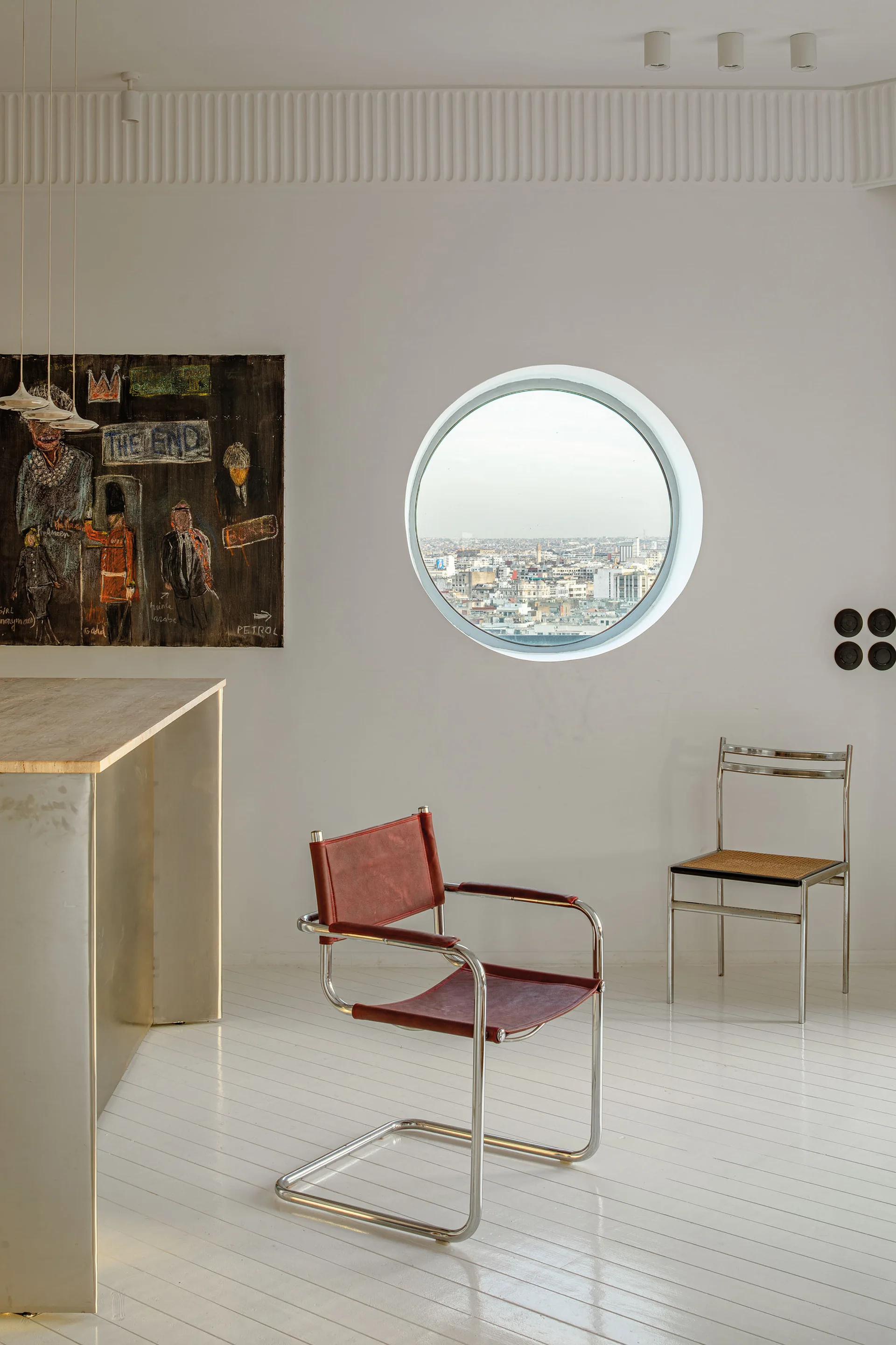 A bright room with white painted wood floors, a round window showing a city view, a red leather armchair, and a metal chair with a cane seat.
