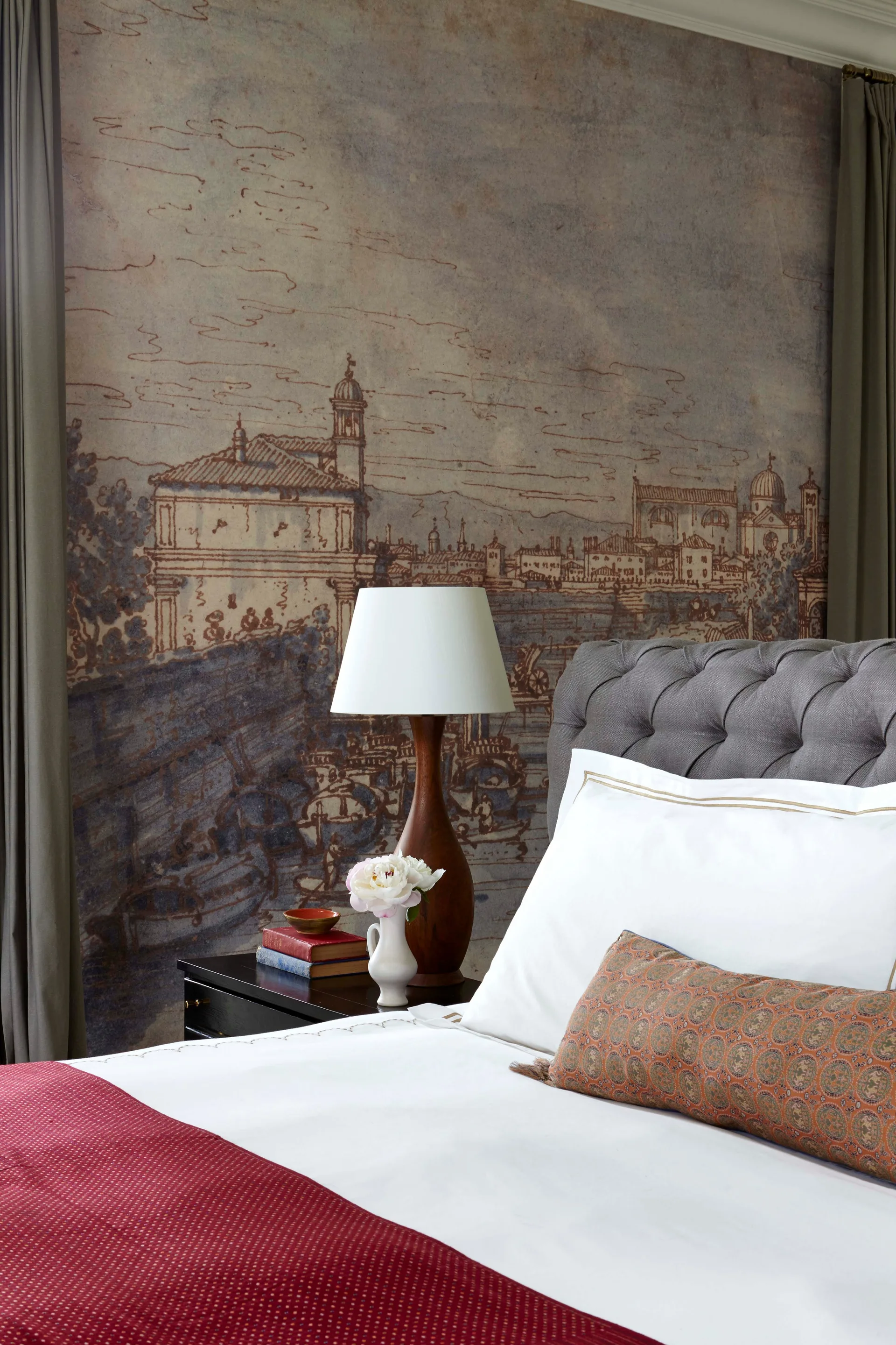 A bedroom features a grey tufted headboard against a sepia-toned cityscape wall mural, flanked by a dark wood nightstand with a lamp.