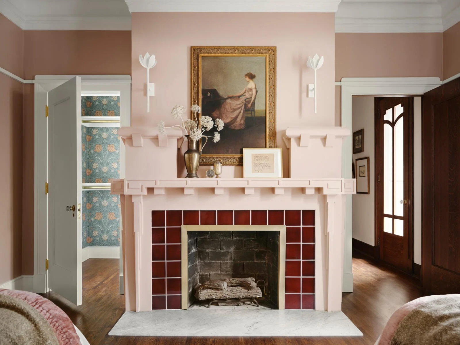 A pink painted fireplace mantel features glossy deep red tiles around the hearth, flanked by white flower sconces and topped with a framed portrait.