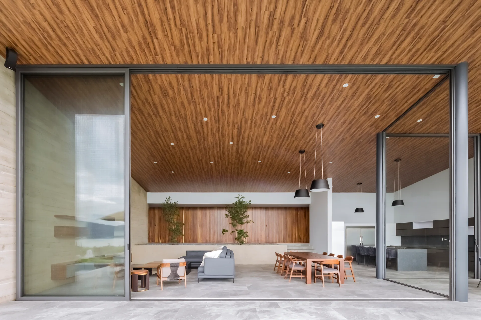A covered modern patio features a warm wood-plank ceiling, large sliding glass doors, a grey sofa seating area, and a wooden dining set.