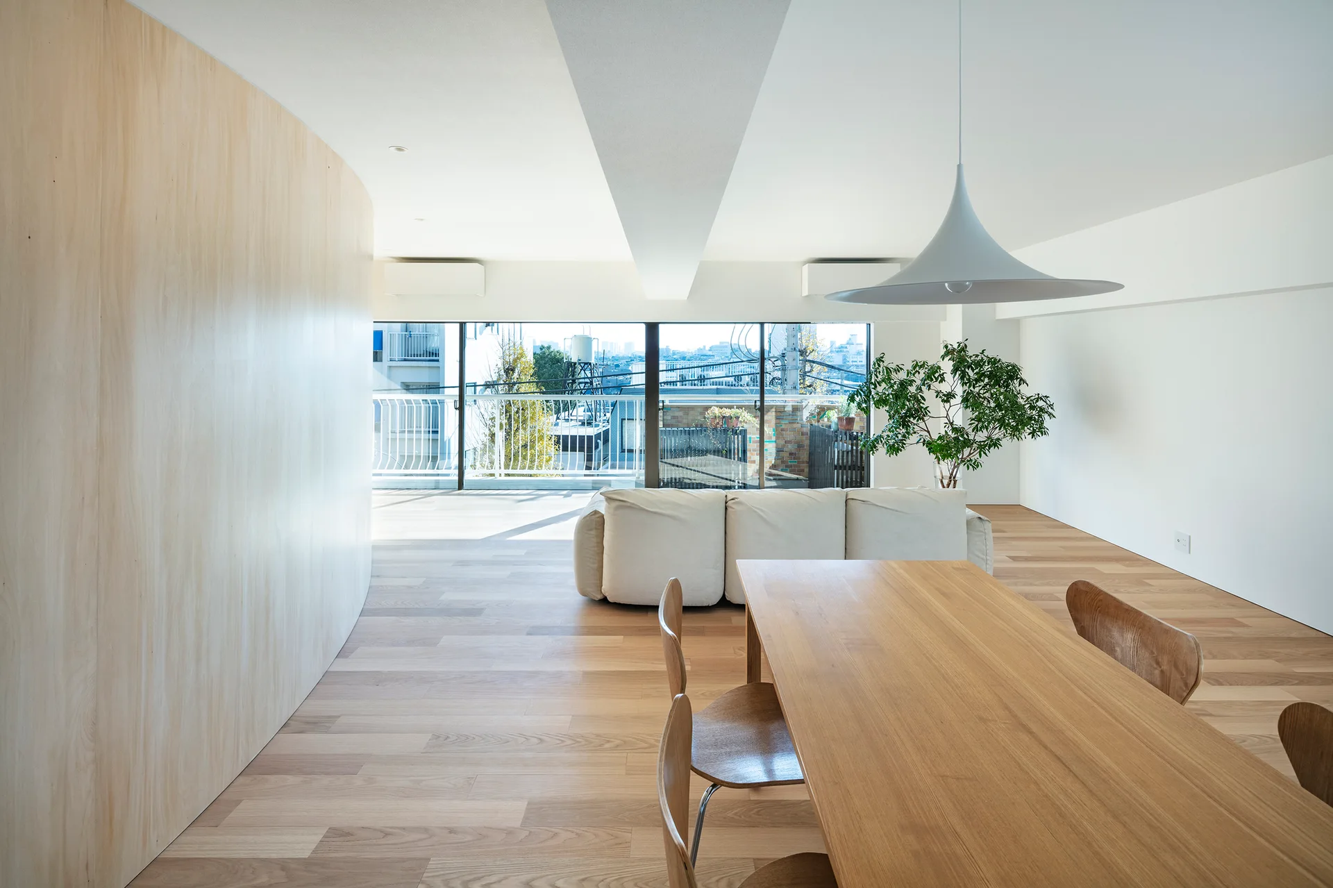 A minimalist open-plan dining and living area features a curved light wood wall, a wooden dining set, a low white sofa, and large windows.