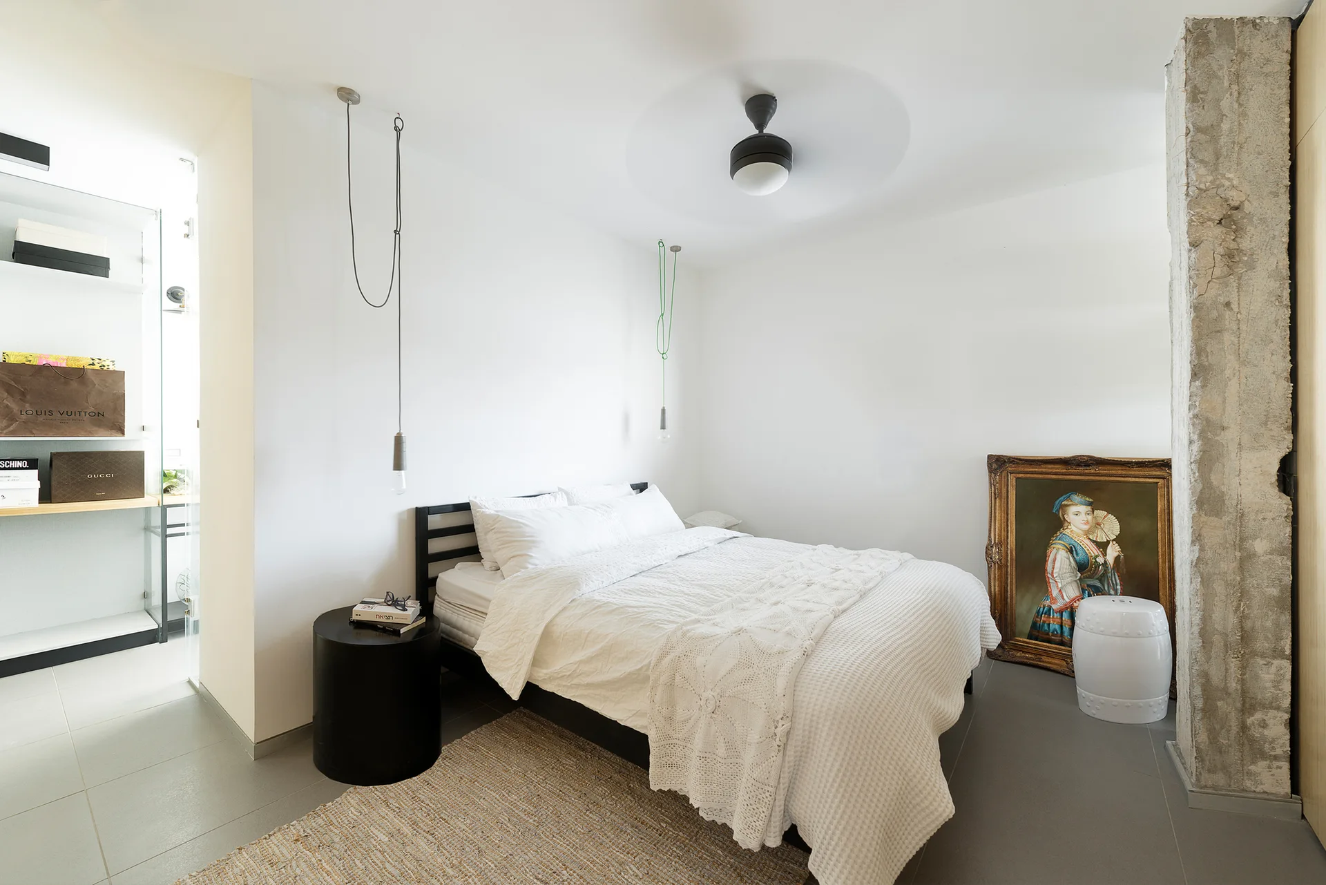 A minimalist bedroom features a bed with white bedding, an exposed concrete pillar, and a framed portrait leaning on the floor.
