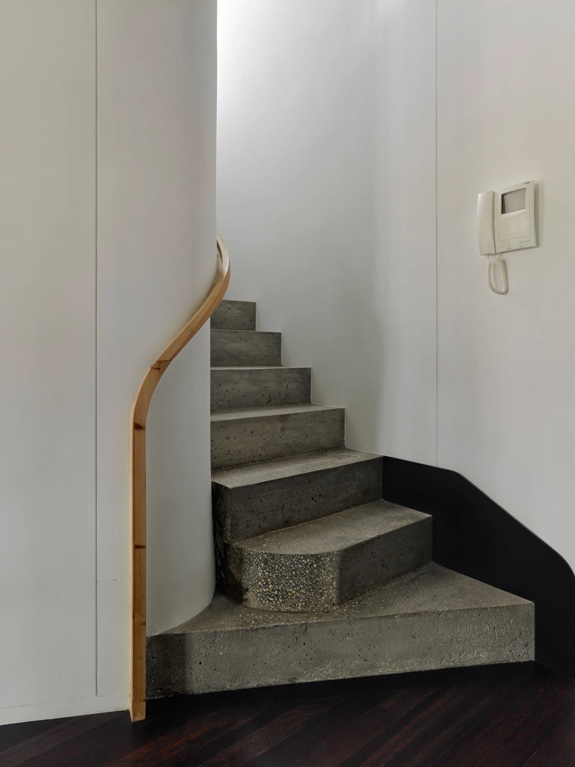 A minimalist concrete staircase with a curved light wood handrail mounted against a rounded white wall.