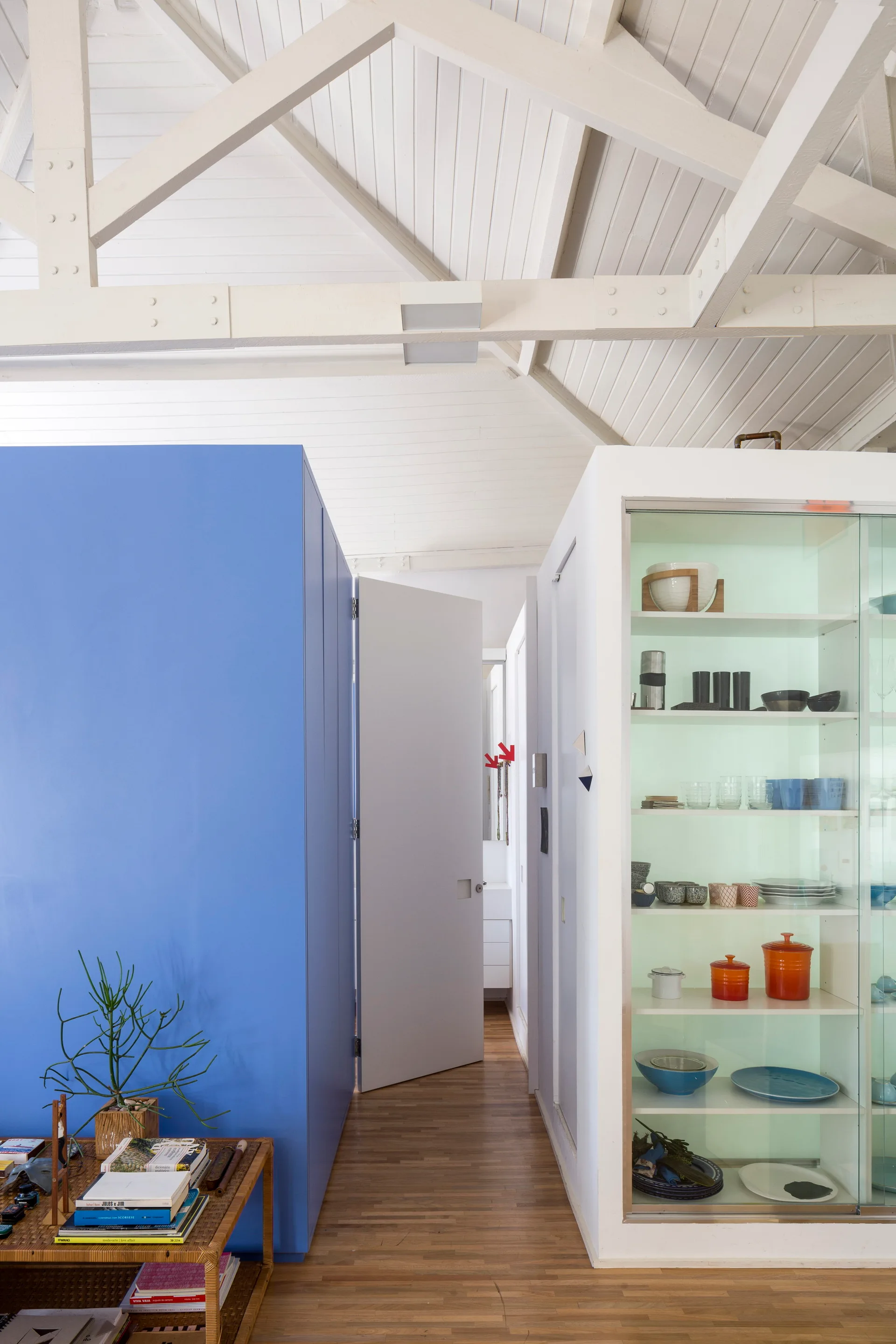 An interior hallway features a bold blue partition wall, a white glass-fronted display cabinet filled with dishes, and exposed white ceiling trusses.