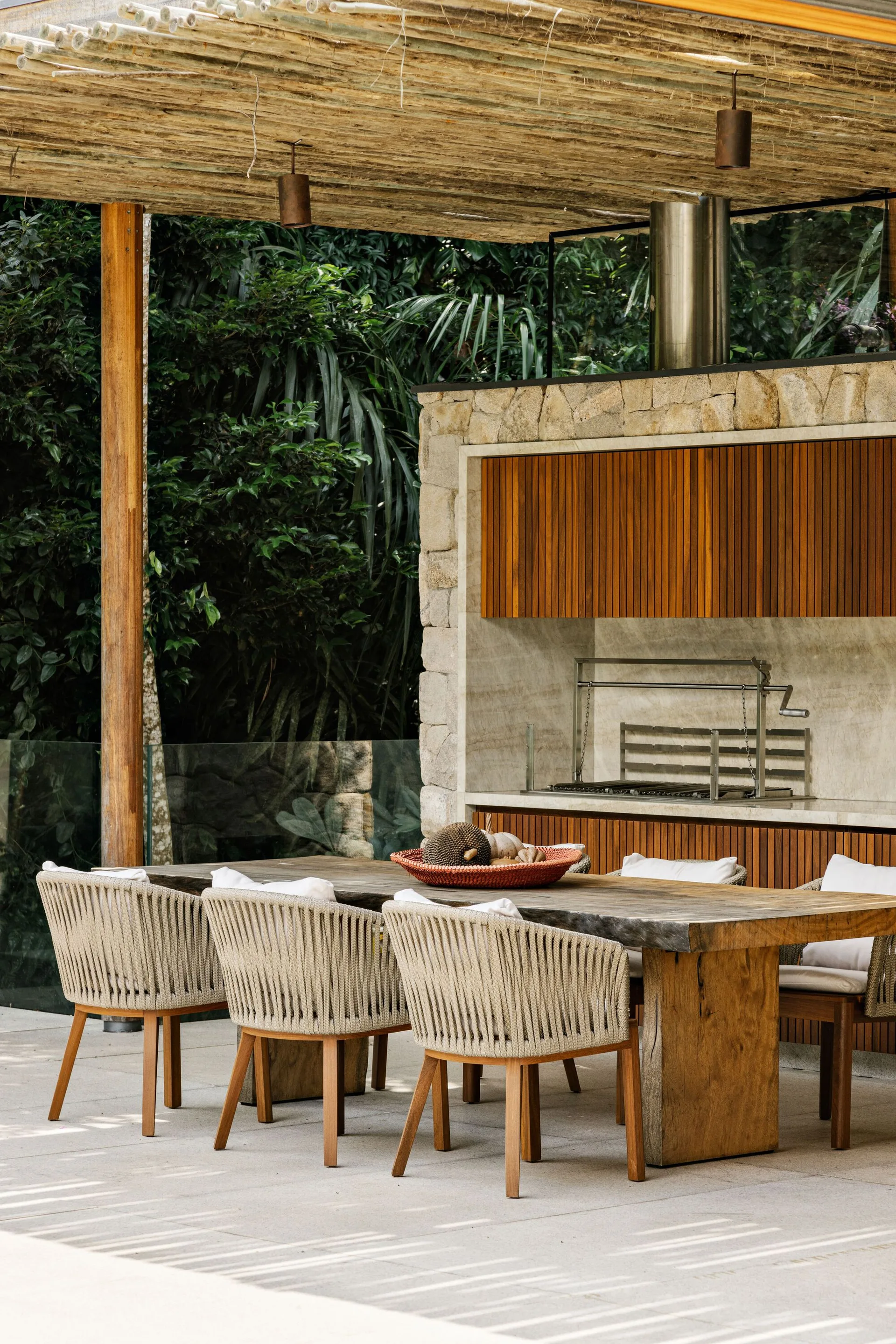 An outdoor dining area featuring a live-edge wooden table, woven rope chairs, and a built-in stone grill station backed by lush green foliage.