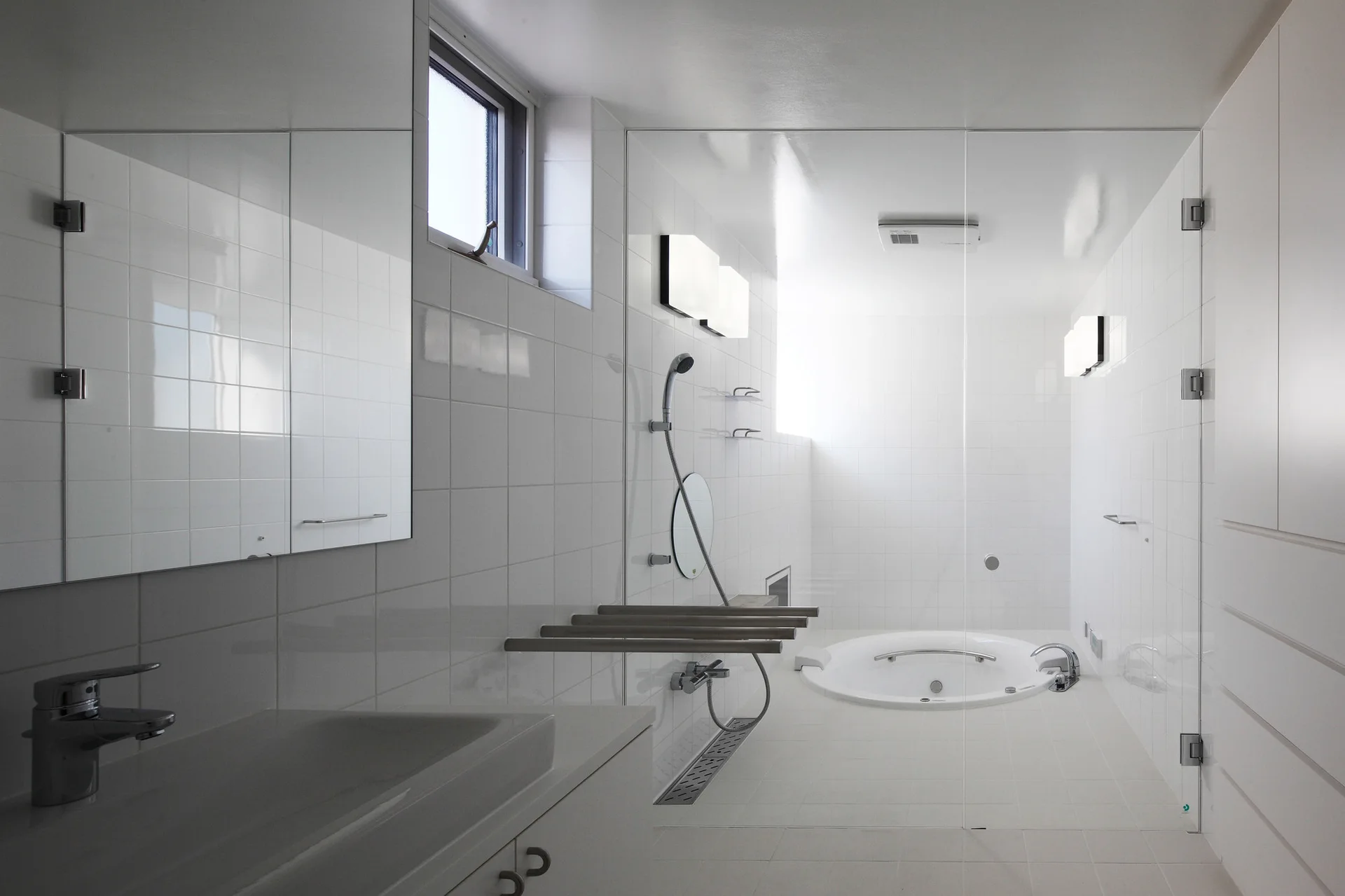 A minimalist white bathroom features square tile walls, a vanity with a mirror, a glass partition, and a sunken circular soaking tub in a wet room.