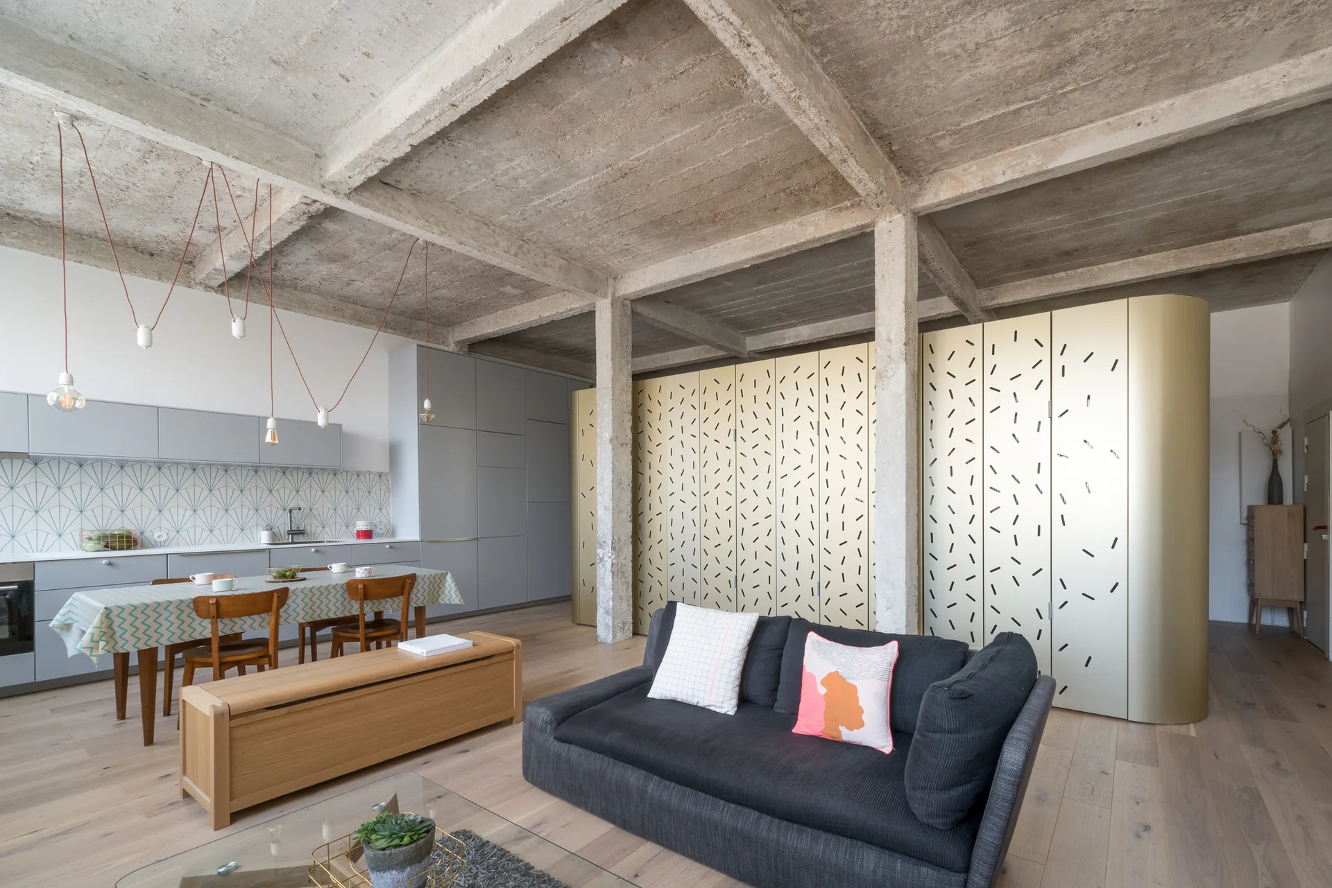 A contemporary open-concept living area and kitchen featuring an exposed concrete ceiling, a grey sofa, and a large patterned gold storage unit.