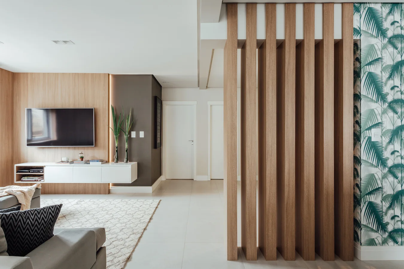 A modern living room features a vertical wood slat partition wall next to botanical wallpaper and a floating white media console.