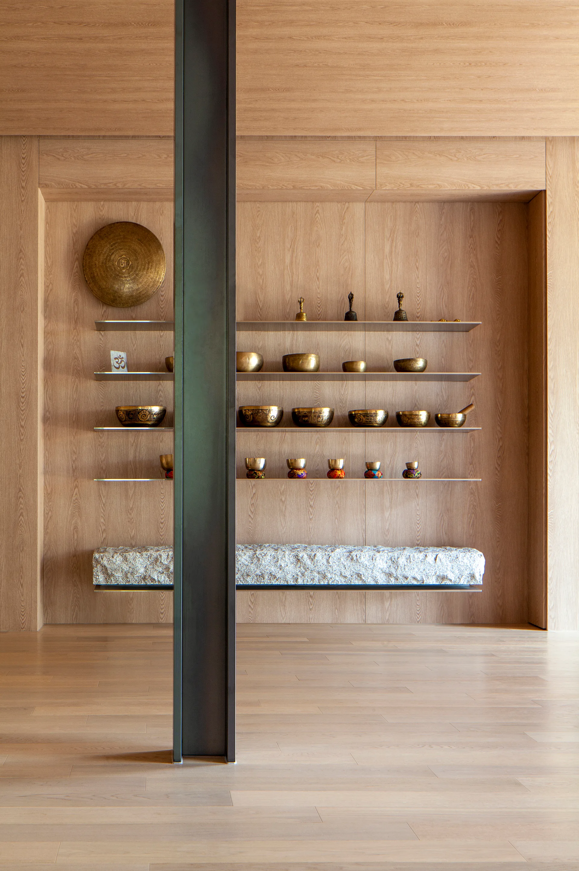 A wood-paneled wellness room features a recessed niche with floating shelves holding singing bowls and a rough stone bench behind a steel column.