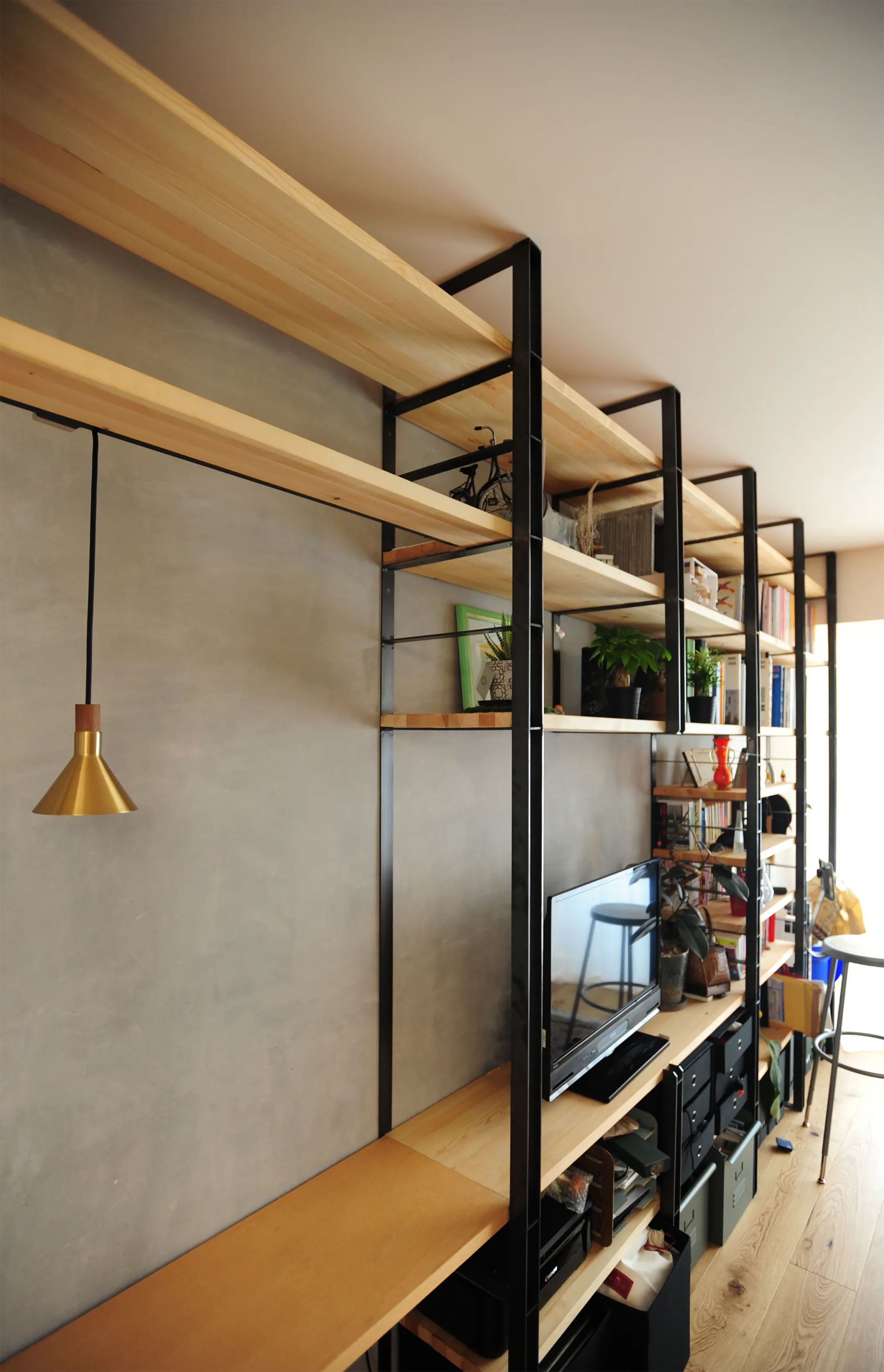 A tall industrial-style shelving unit with black metal frames and light wood planks stands against a textured grey concrete wall.