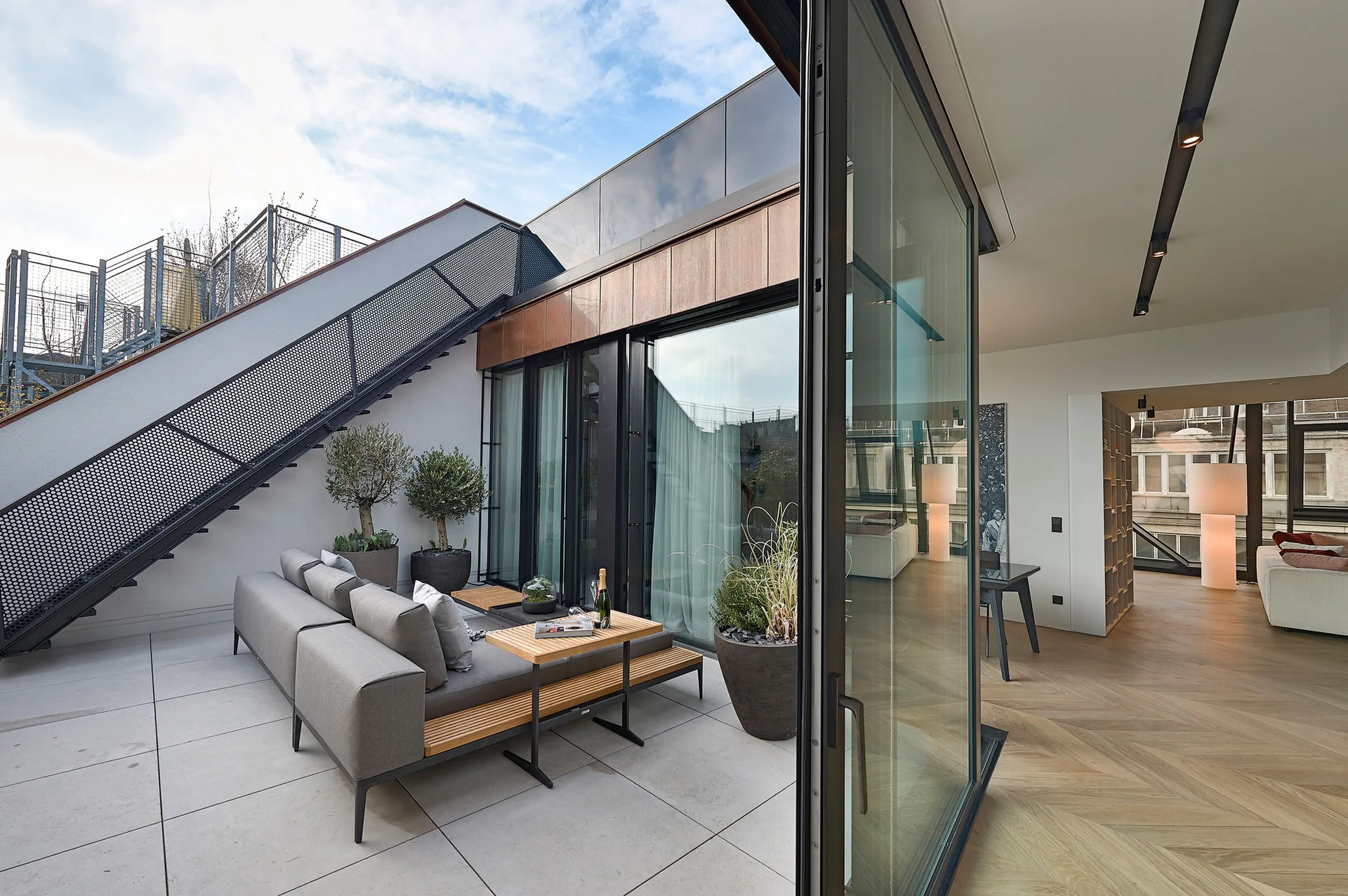 A modern terrace featuring a grey sectional sofa with integrated wooden tables, next to a metal staircase and large sliding glass doors leading indoors.