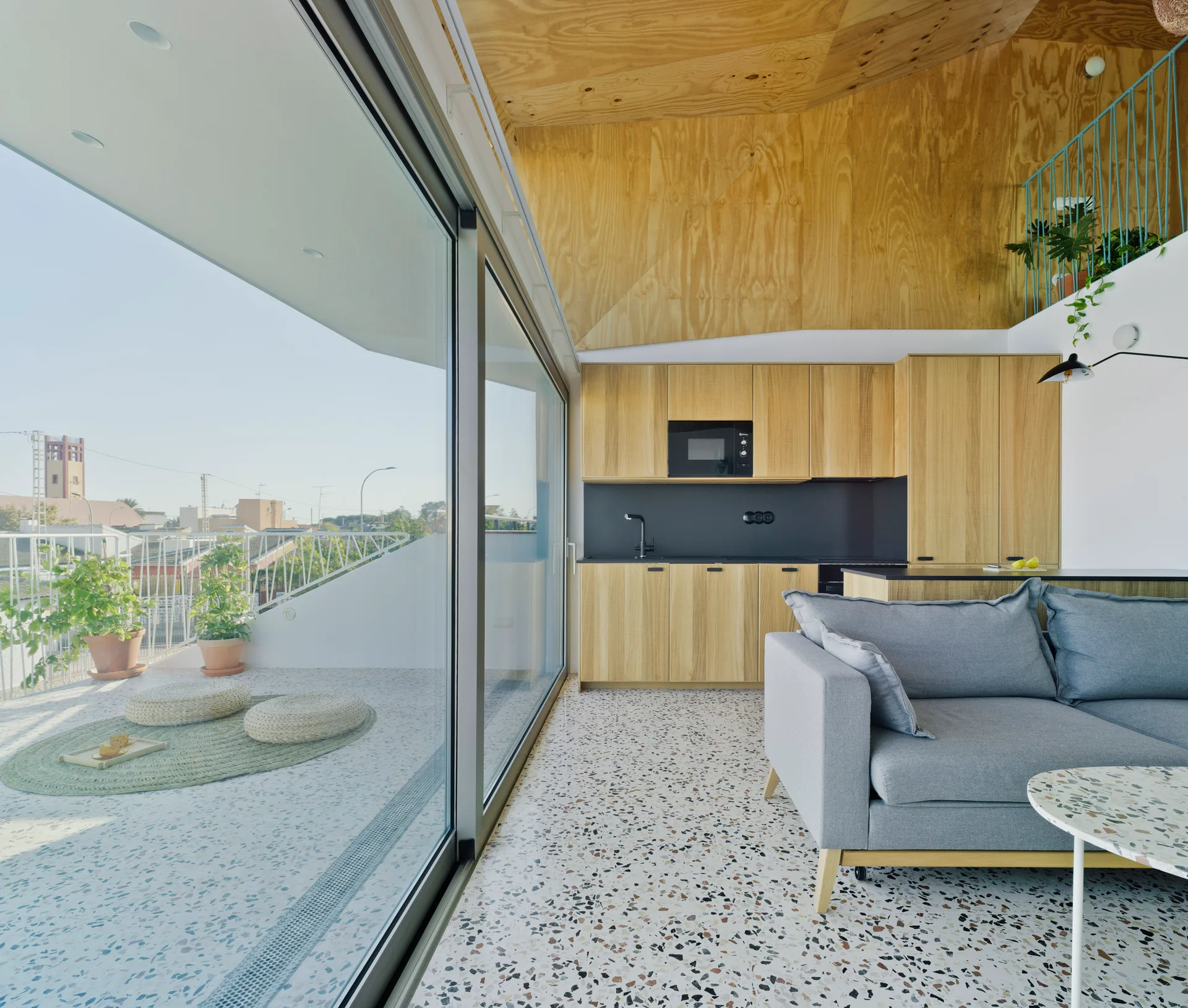 A modern living area features a grey sofa, light wood kitchen cabinets, speckled terrazzo flooring, and a large sliding glass door opening to a balcony.