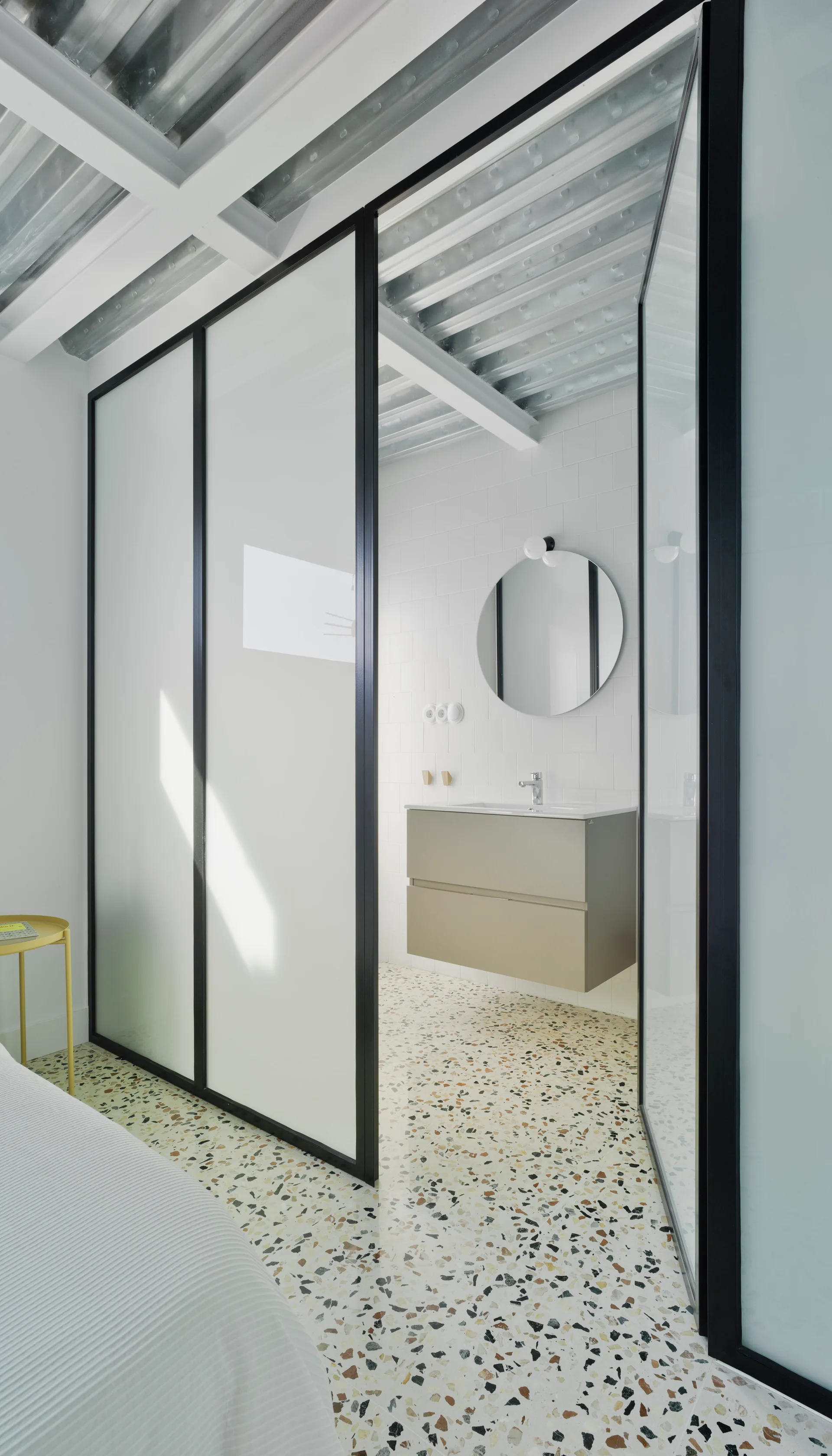 A modern bathroom with a floating beige vanity, round mirror, and white tiled walls, separated from a bedroom by a black-framed glass partition.