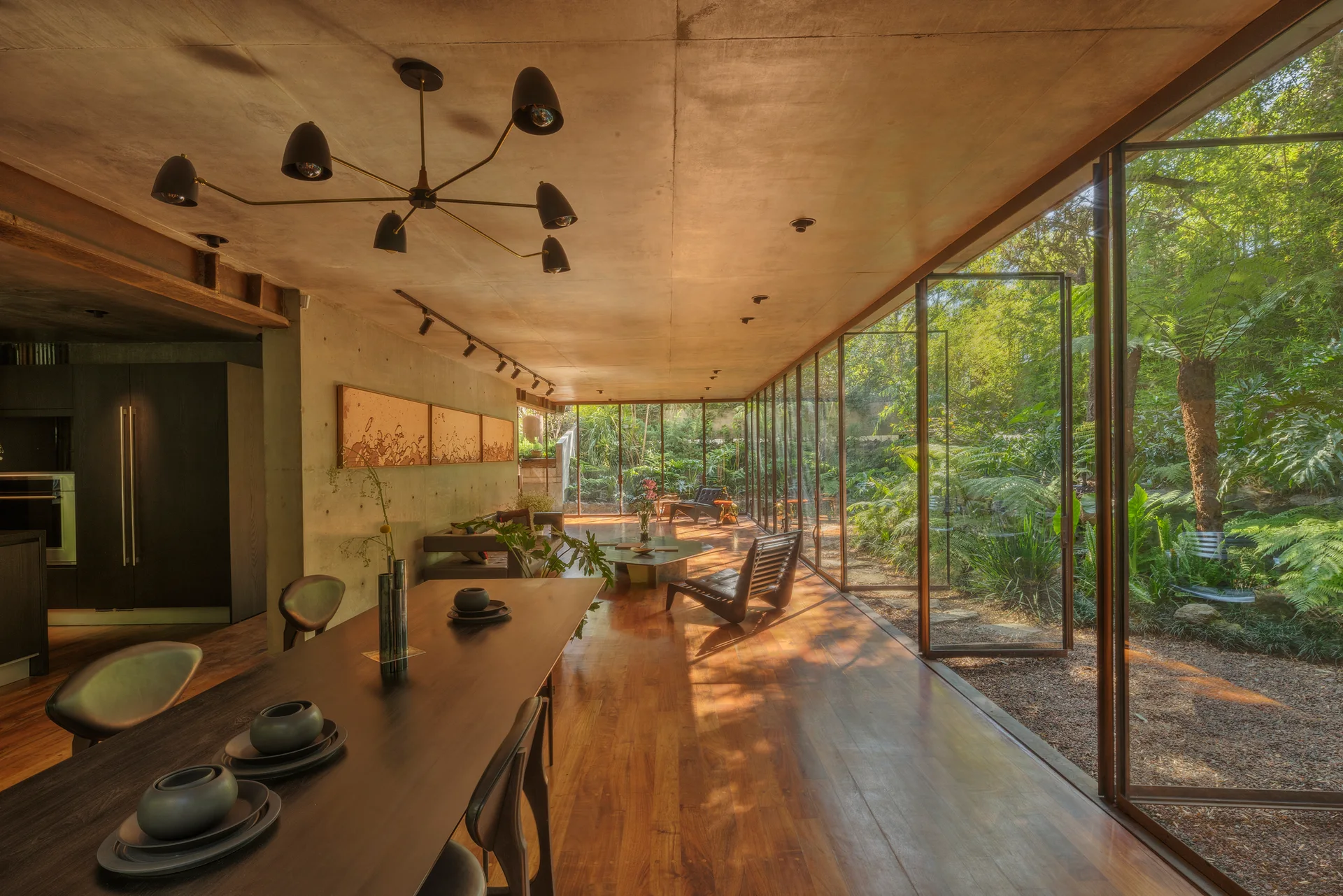 An open dining and living space features a long dark wood table, exposed concrete walls, and floor-to-ceiling pivoting glass doors opening to a lush garden.