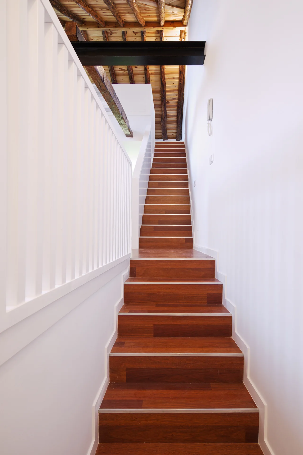A wooden staircase with rich brown treads ascends between white walls under a ceiling with exposed rustic timber beams and a black steel support.