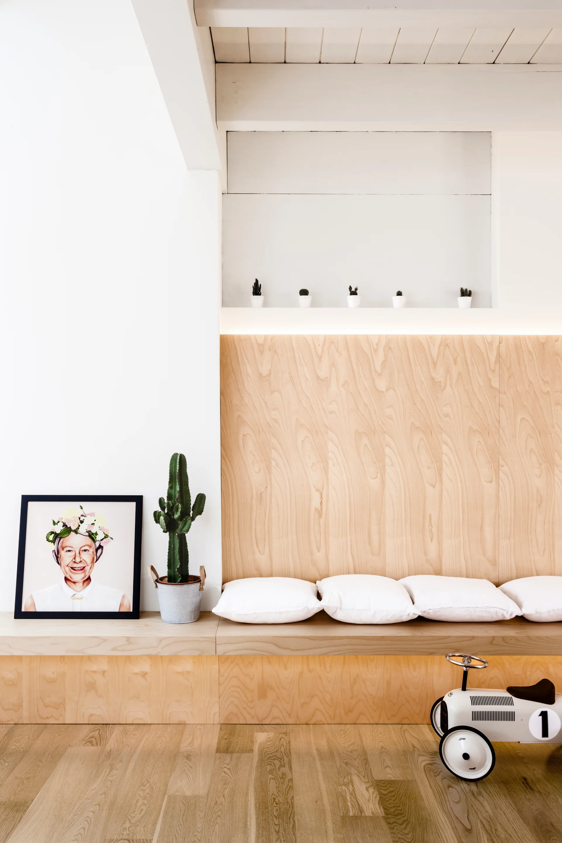 A light wood built-in bench with white cushions features matching wood back paneling, a recessed wall shelf with small cacti, and a white toy car nearby.