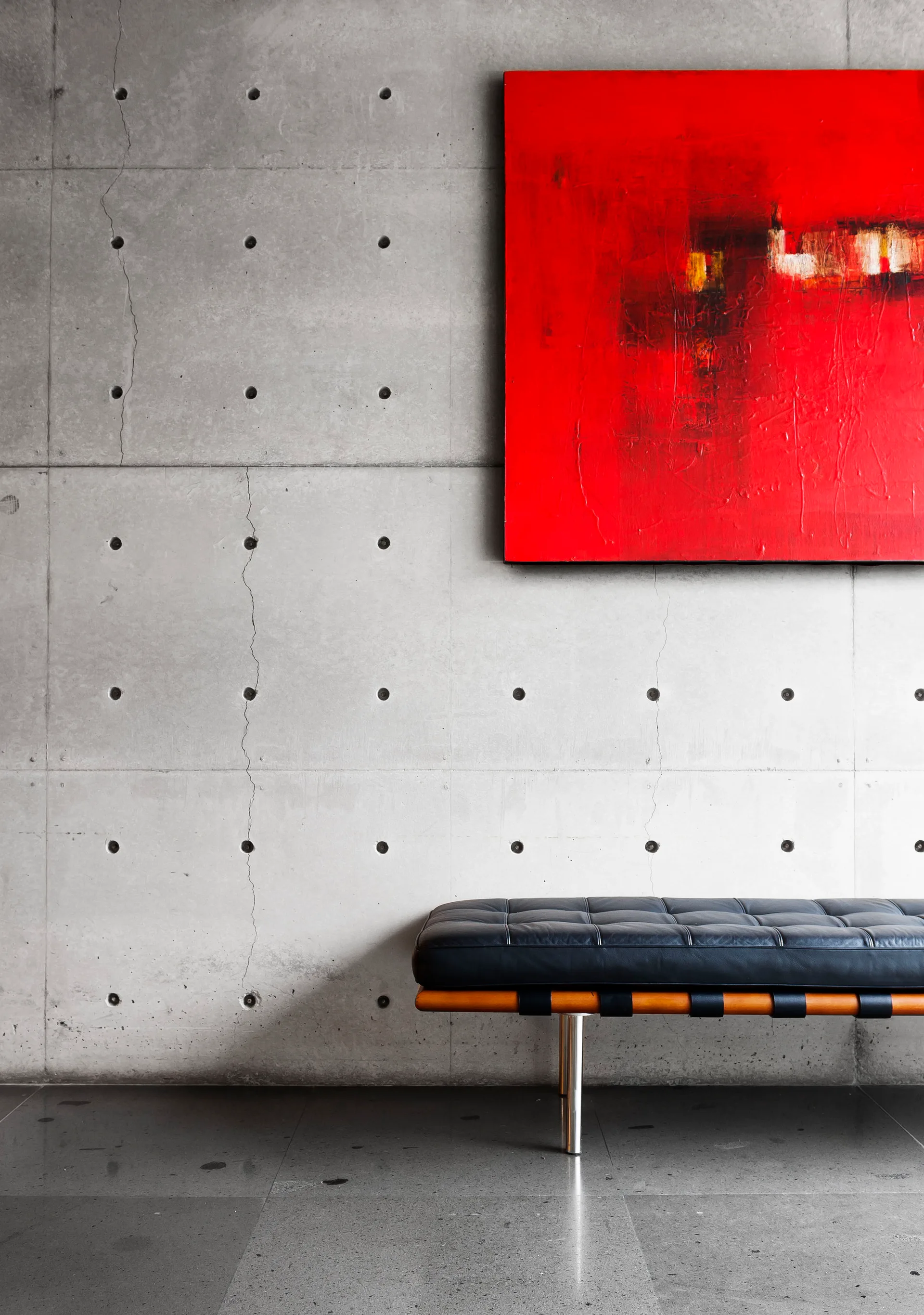 A tufted black leather bench sits against a grey concrete wall featuring circular tie holes, positioned beneath a large red abstract painting.