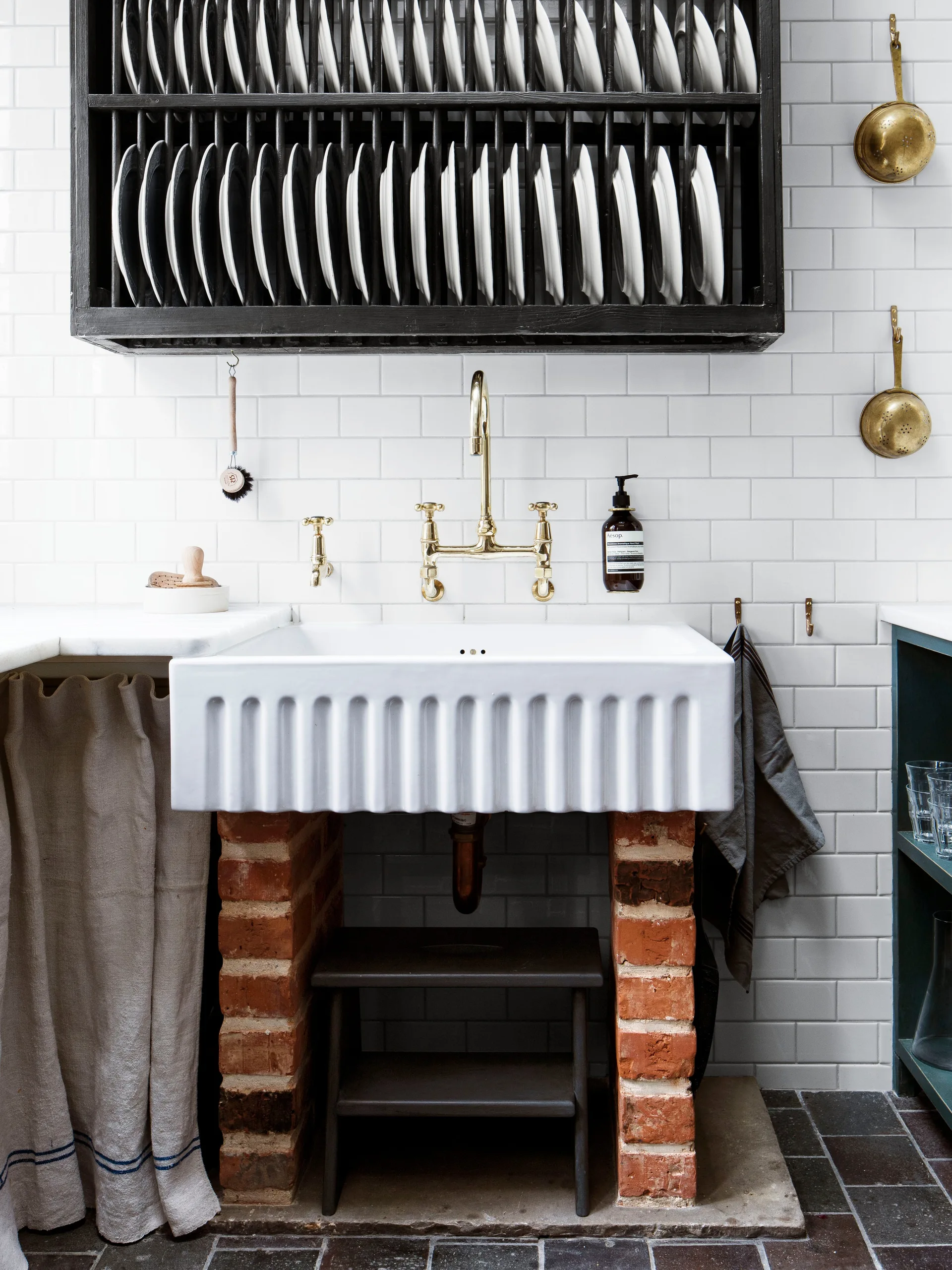 A white fluted farmhouse sink sits on exposed brick supports against a white subway tile wall with a black wooden plate rack hanging above.