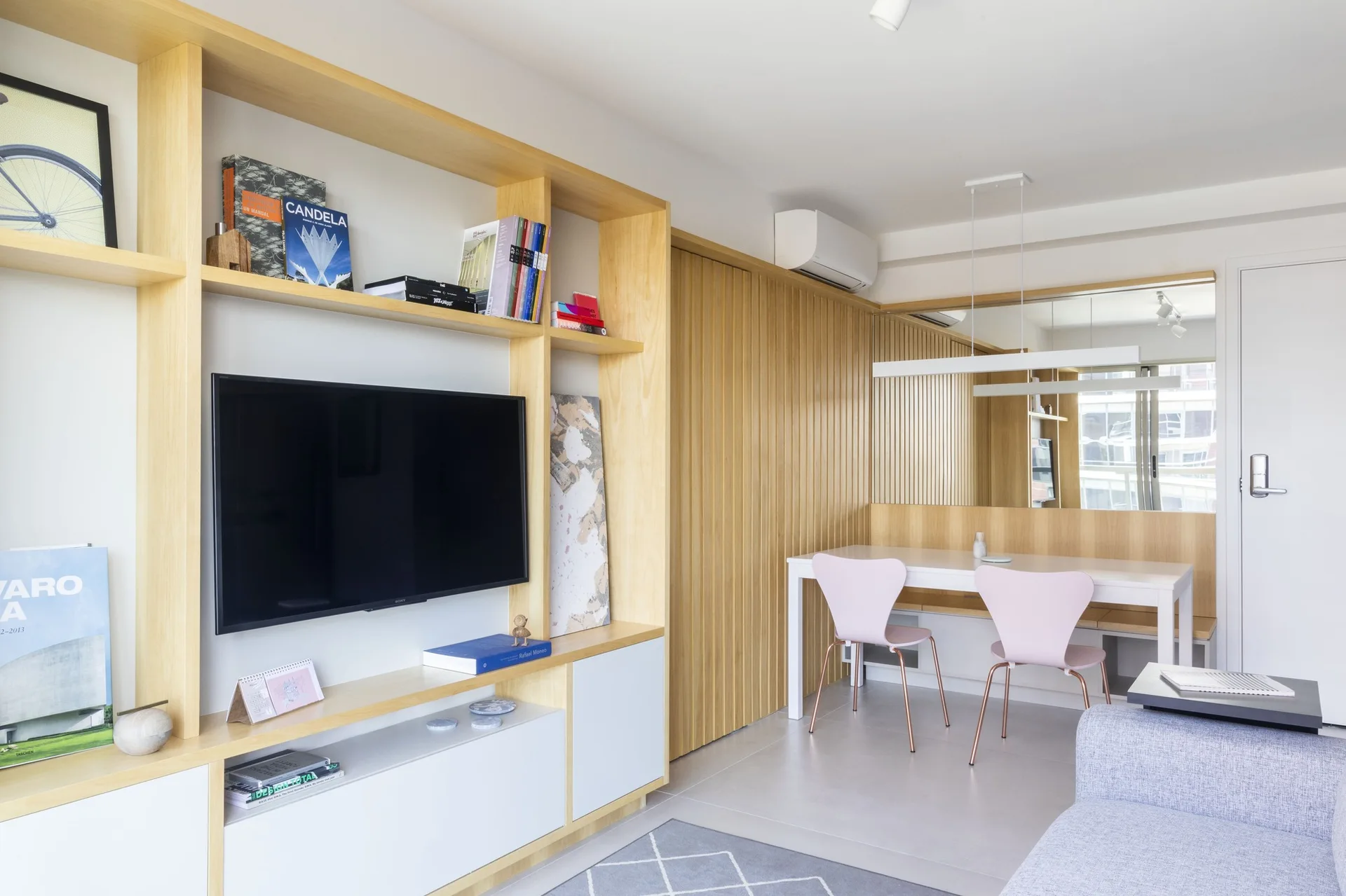 A modern living area featuring a light wood built-in media unit with a TV, a vertical slat wood wall, and a dining table with pink chairs.