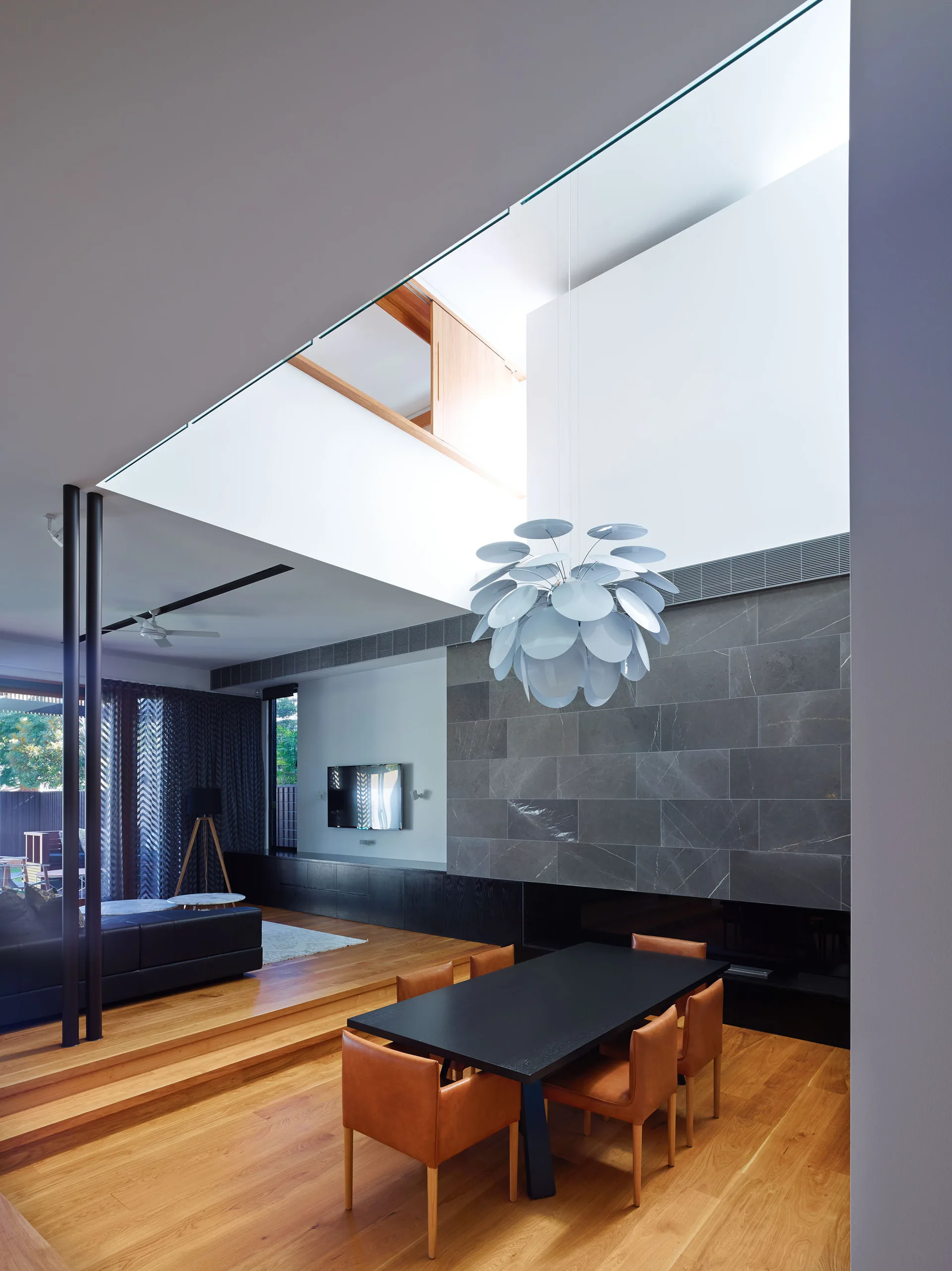 A split-level dining area features a black dining table with tan leather chairs below a large skylight and a grey stone fireplace wall.