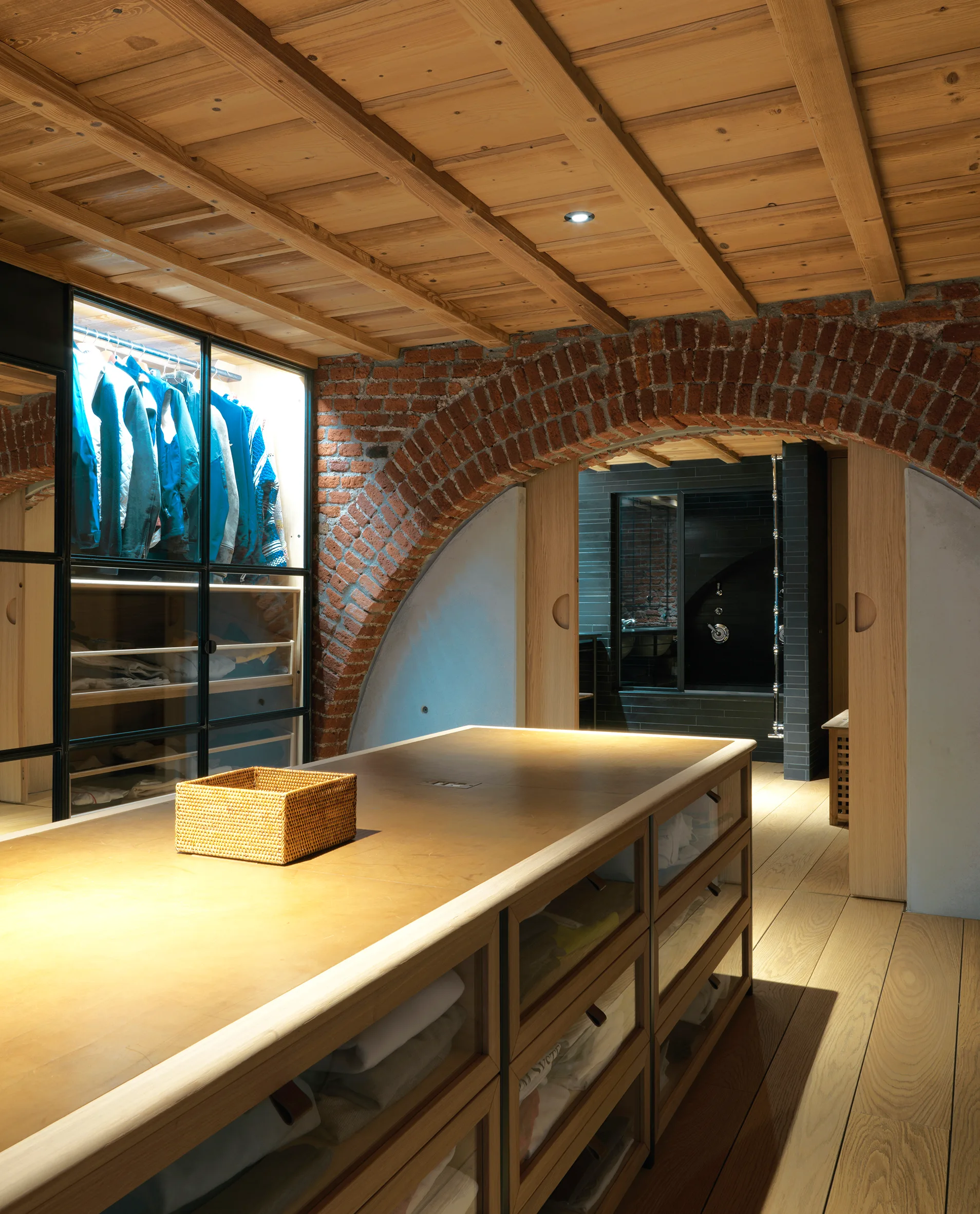 A walk-in dressing room features a large wood storage island, glass-fronted backlit closets, and an exposed brick archway under a wood plank ceiling.