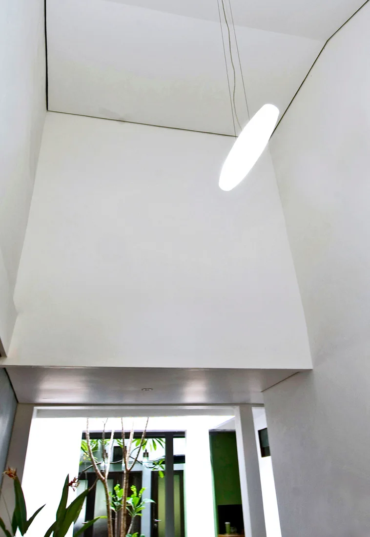 A tall white minimalist entryway features a high ceiling with a suspended glowing disc pendant light and a view of an outdoor courtyard with green plants.