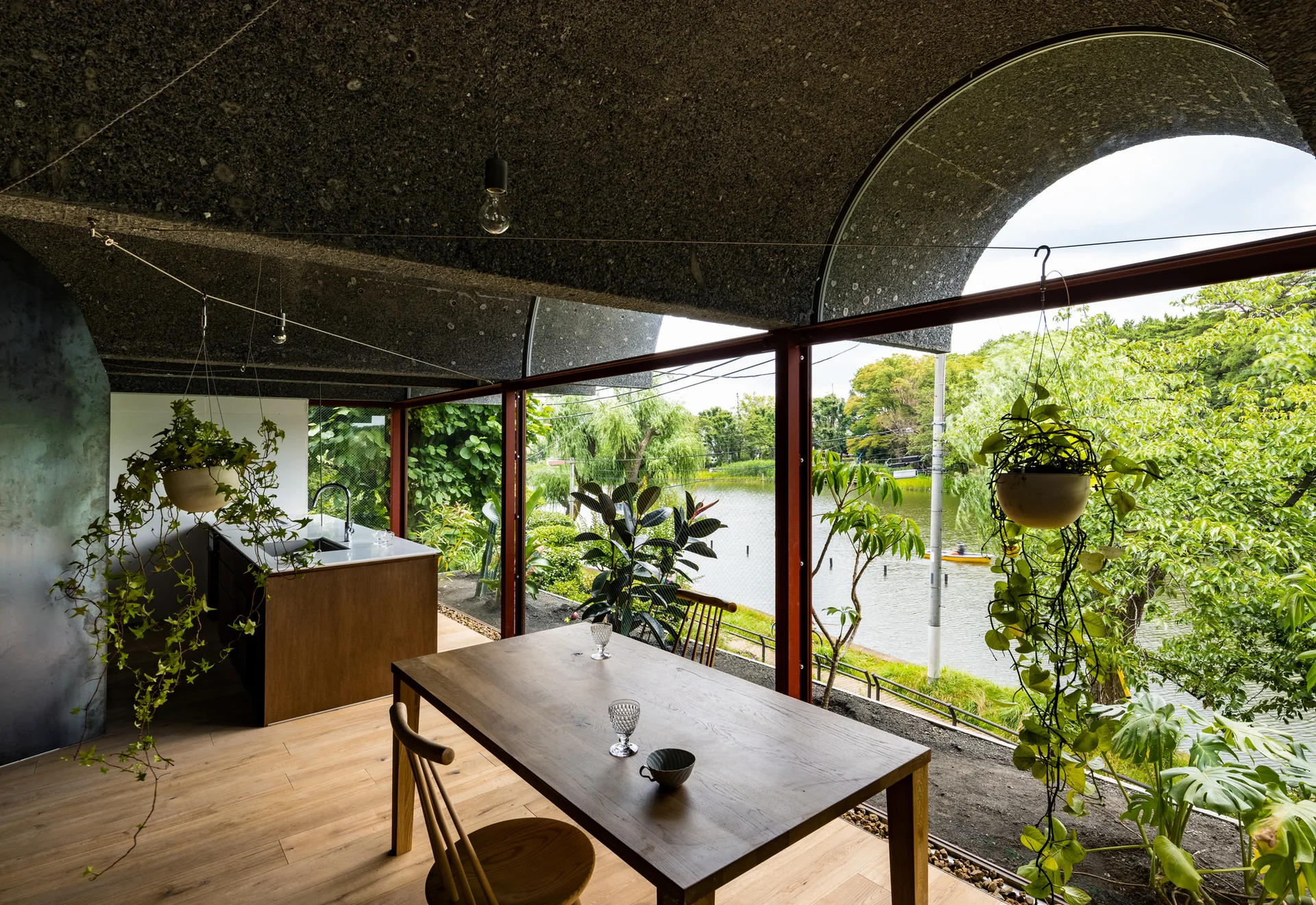 A modern dining area features a wooden table and kitchen island beneath a curved concrete ceiling, with large windows overlooking a scenic lake.