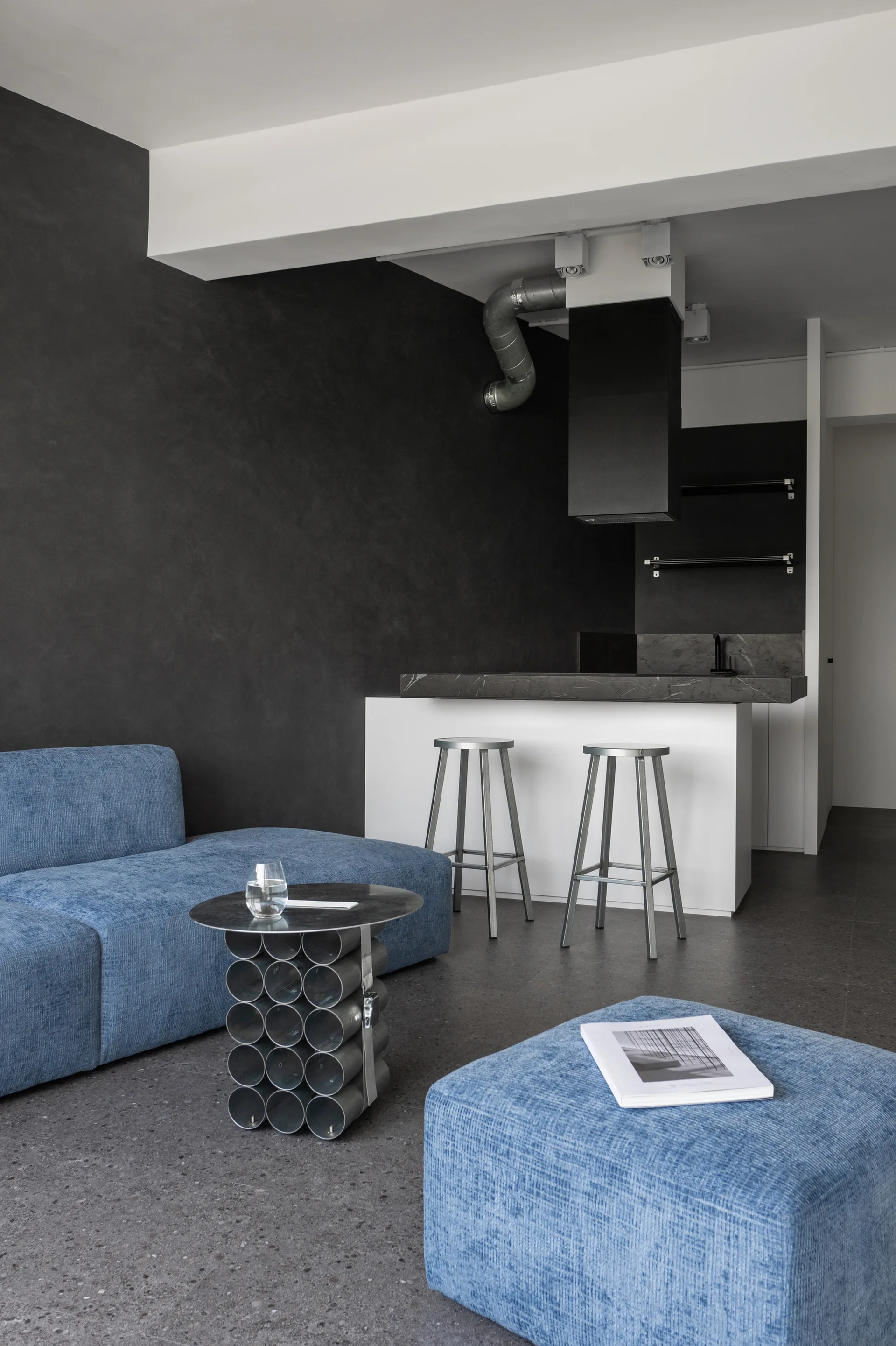 A modern open-plan living space features a blue textured sofa and matching ottoman opposite a white kitchen island with two metal bar stools.