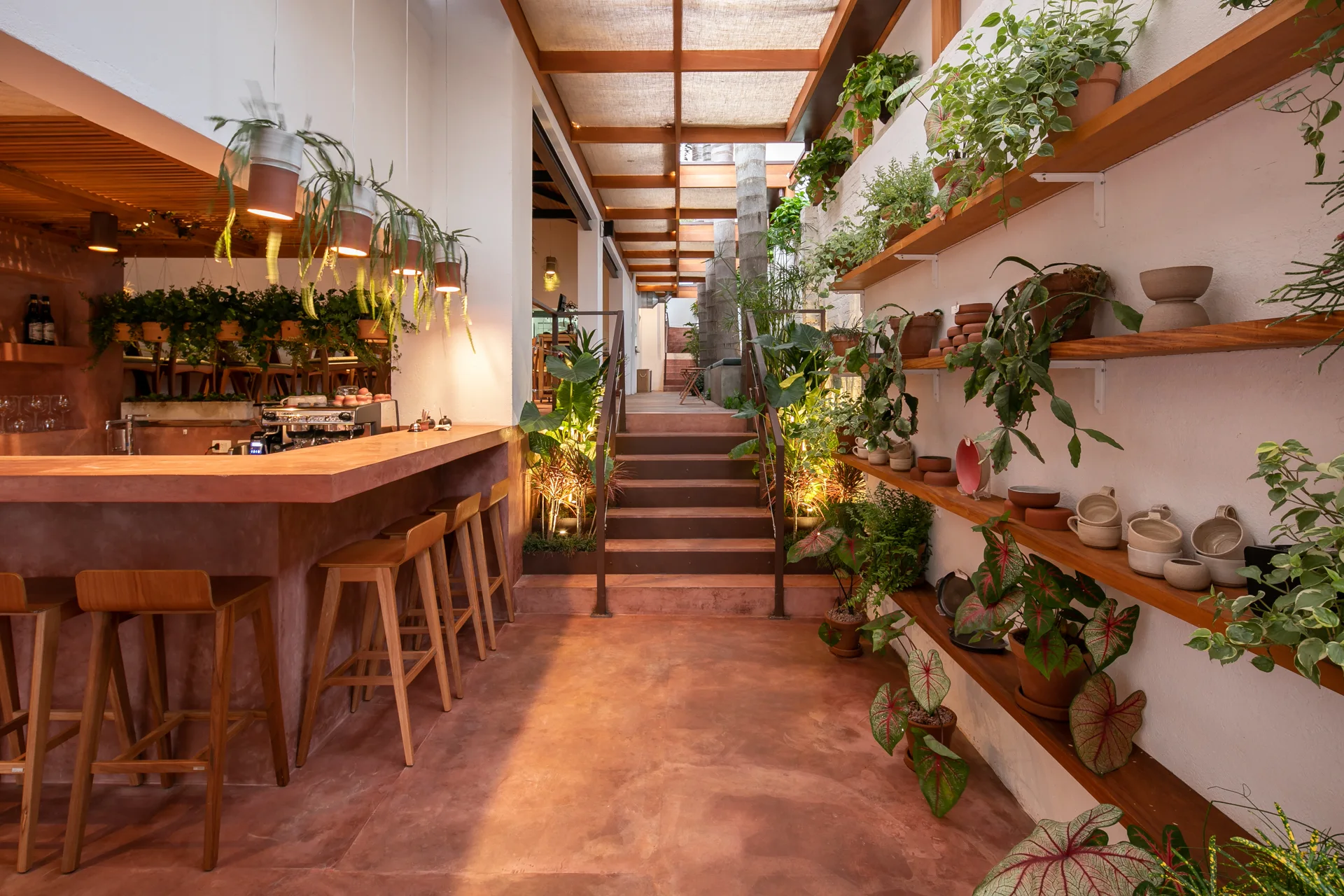 A terracotta-toned bar featuring wooden stools, a central staircase, and a wall of open wood shelves filled with potted plants and ceramics.