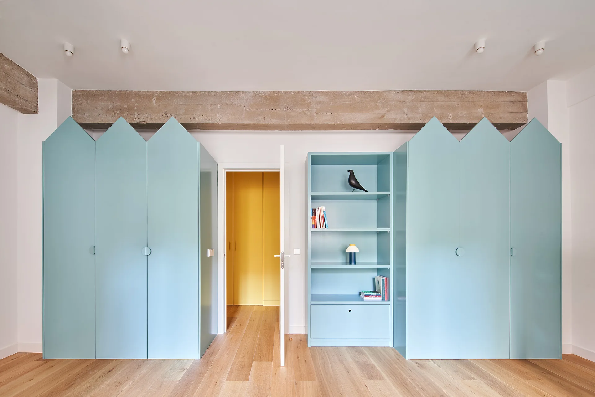Pale blue wardrobes with gabled tops stand on either side of a doorway and open bookshelf beneath an exposed concrete ceiling beam.