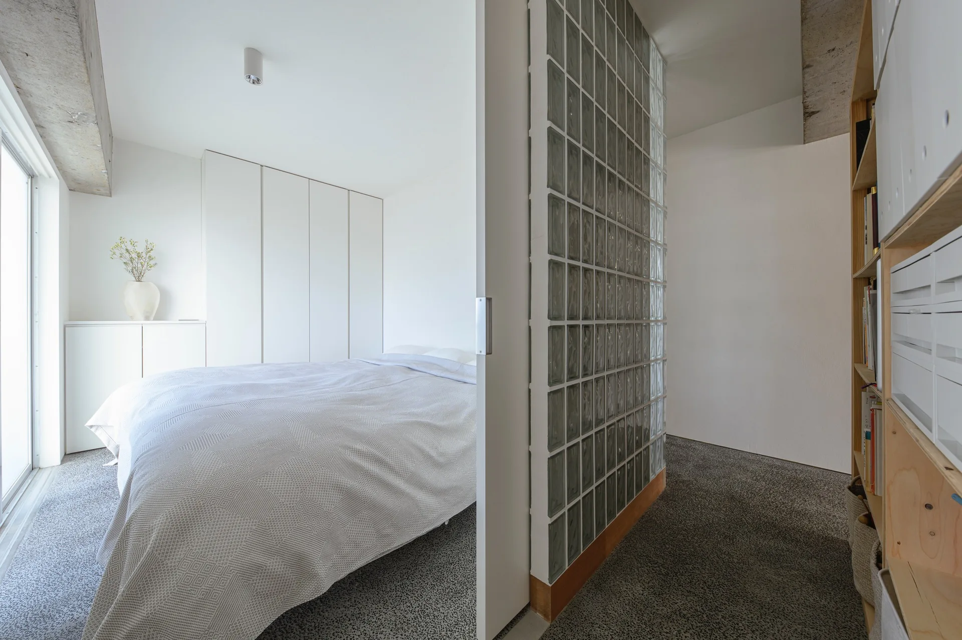 A minimalist bedroom with a bed with patterned gray bedding, a glass block partition wall, and white built-in cabinets.