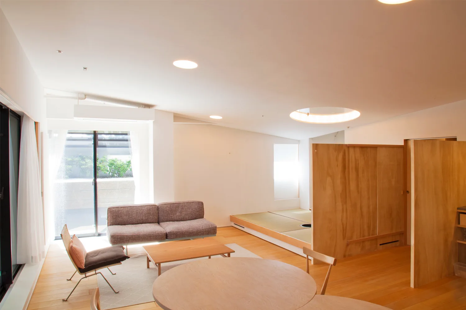 A bright minimalist living room features a round wooden dining table, grey sofa, a tall plywood partition, and a raised tatami platform.