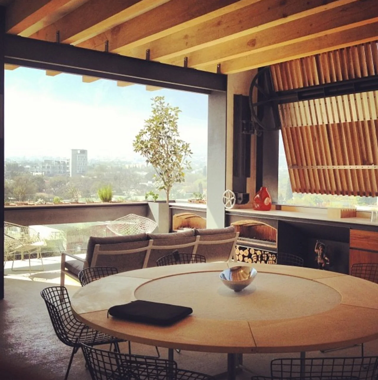 An open-air terrace features a round wood dining table with wire chairs, a grey sofa, exposed wood ceiling beams, and a view of the city skyline.