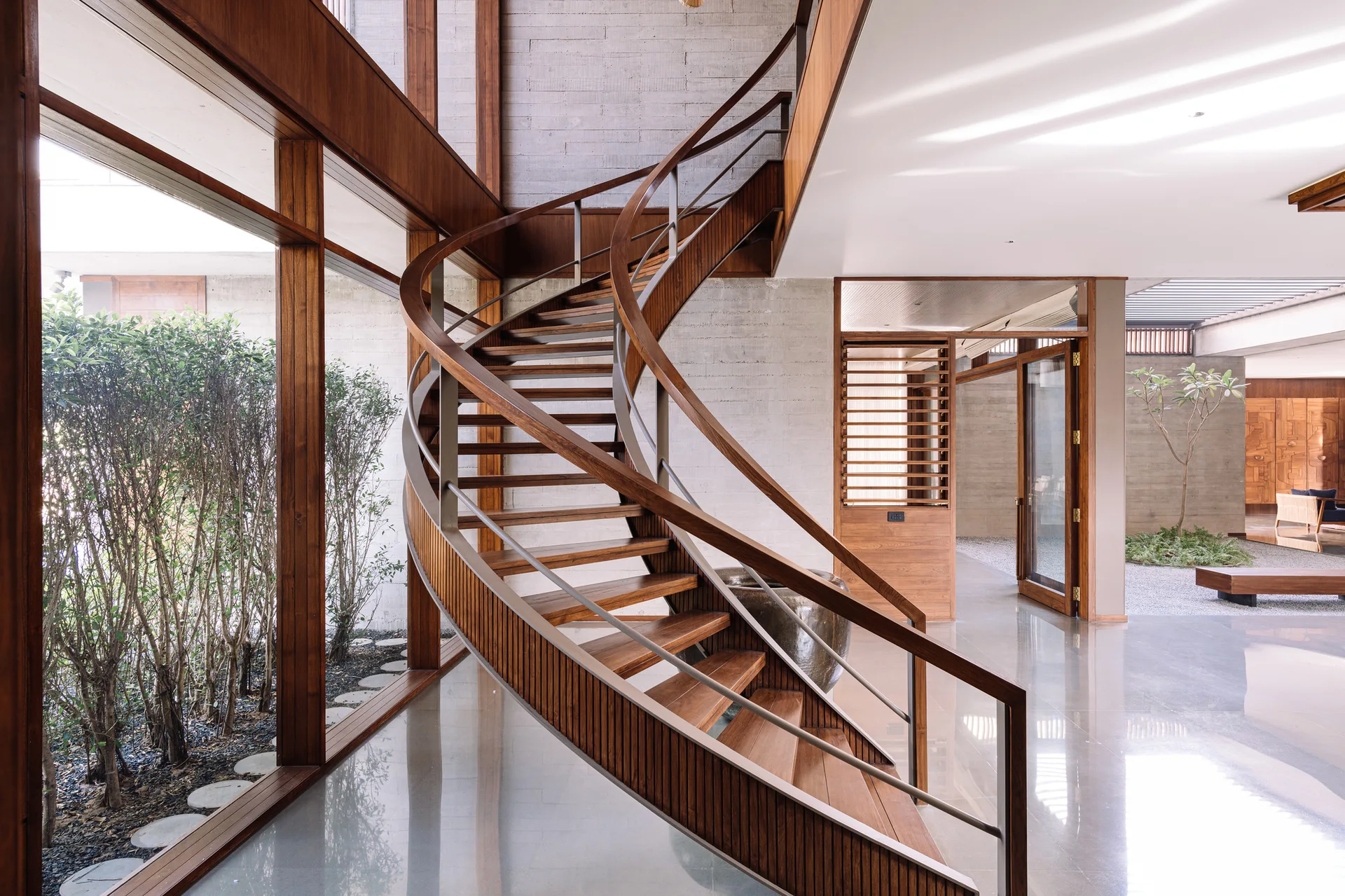 A central curved wooden spiral staircase stands next to floor-to-ceiling glass windows and concrete walls on polished grey floors.