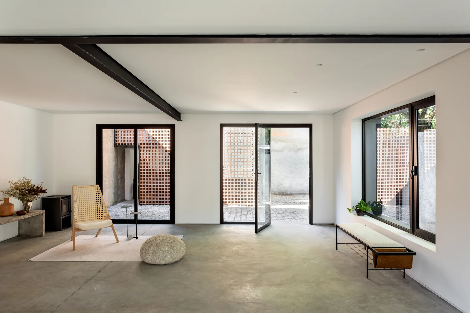 A minimalist living room features a grey concrete floor, a cane armchair on a small rug, white walls, and black-framed glass doors leading outside.
