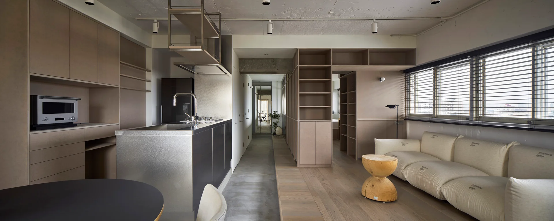 A modern open-plan apartment featuring a cream modular sofa, a brushed metal kitchen island, and extensive built-in wooden shelving units.